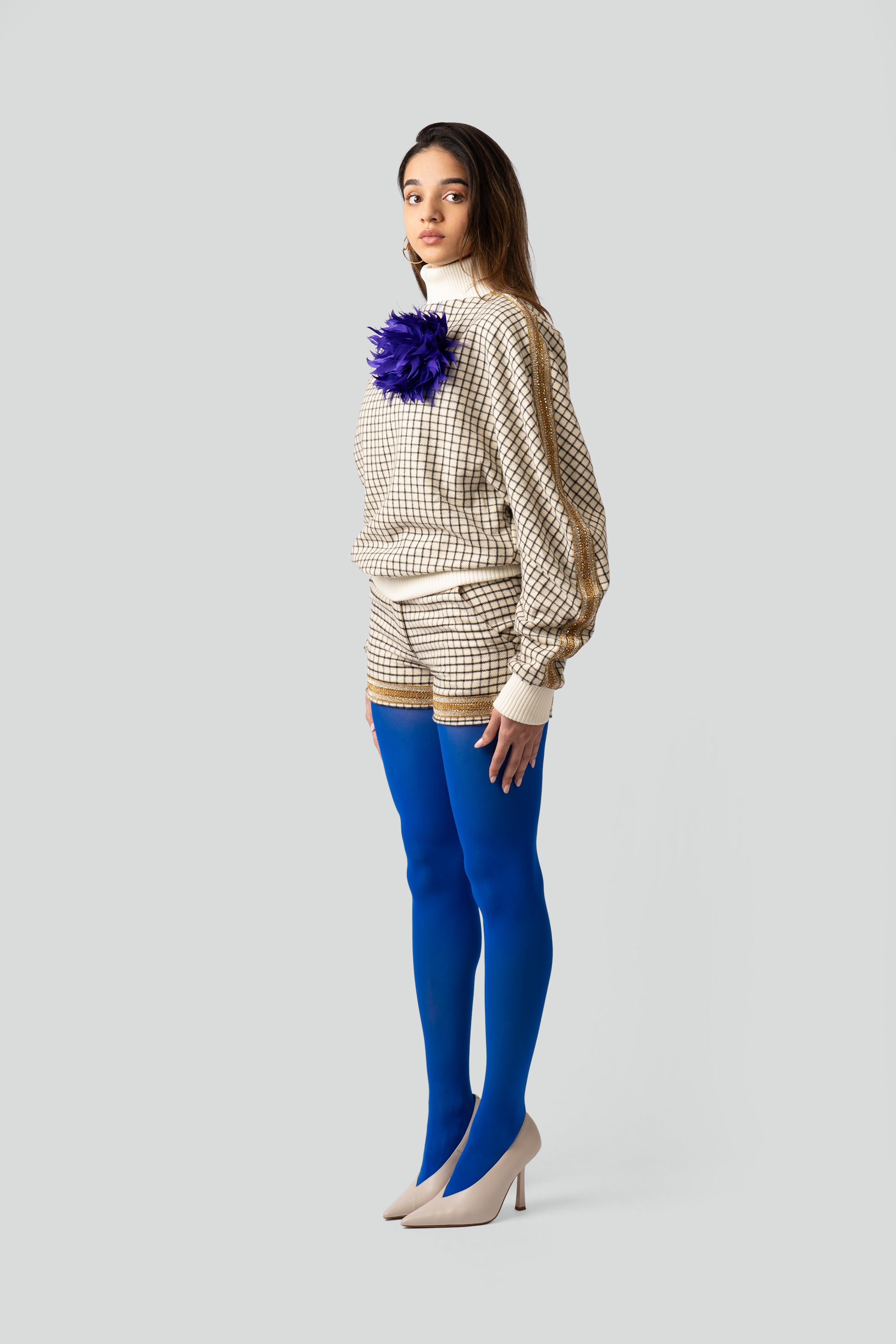 Woman wearing a grid patterned outfit with gold trim and an indigo feathered brooch on a white background