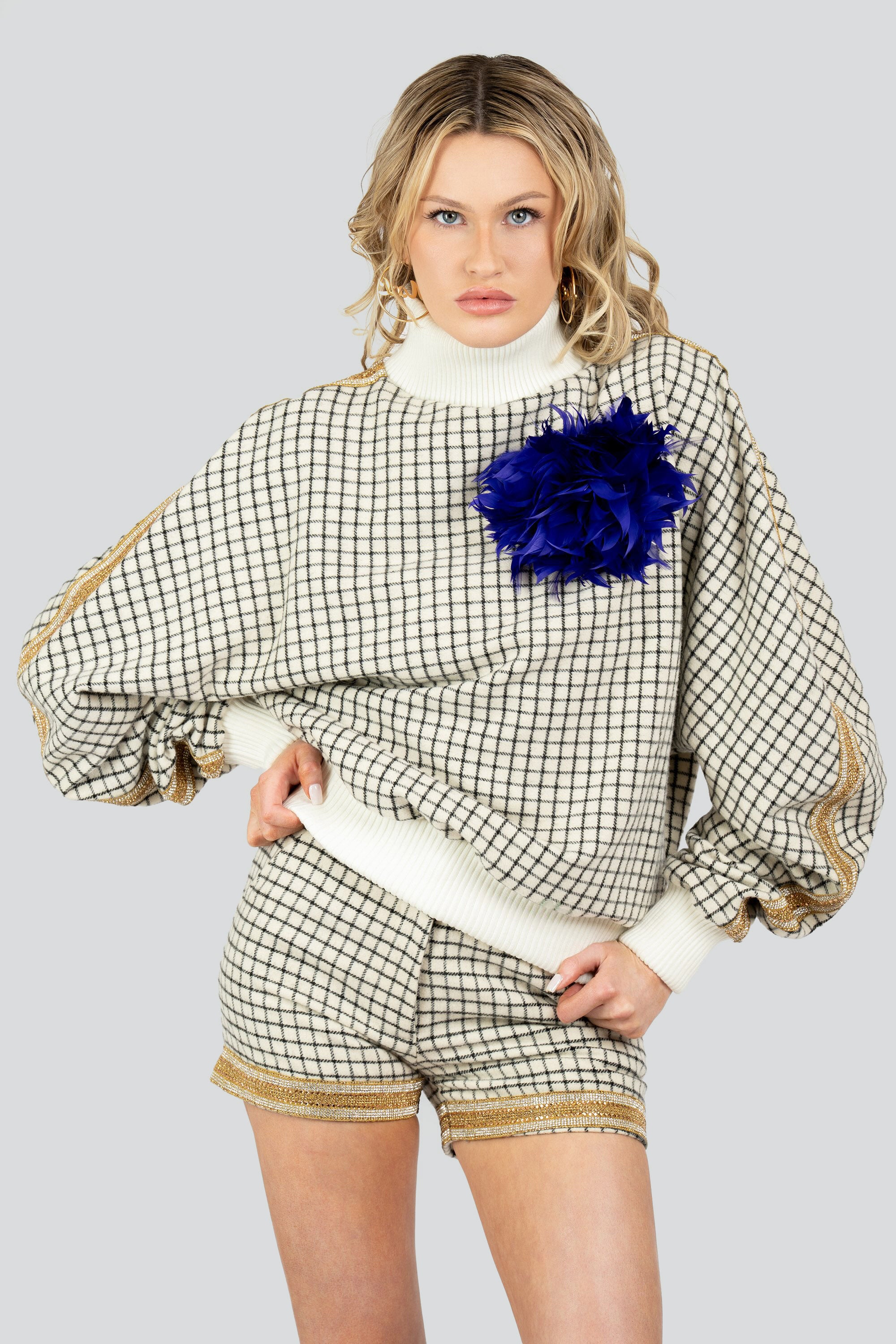 Woman wearing a grid patterned outfit with gold trim and an indigo feathered brooch on a white background