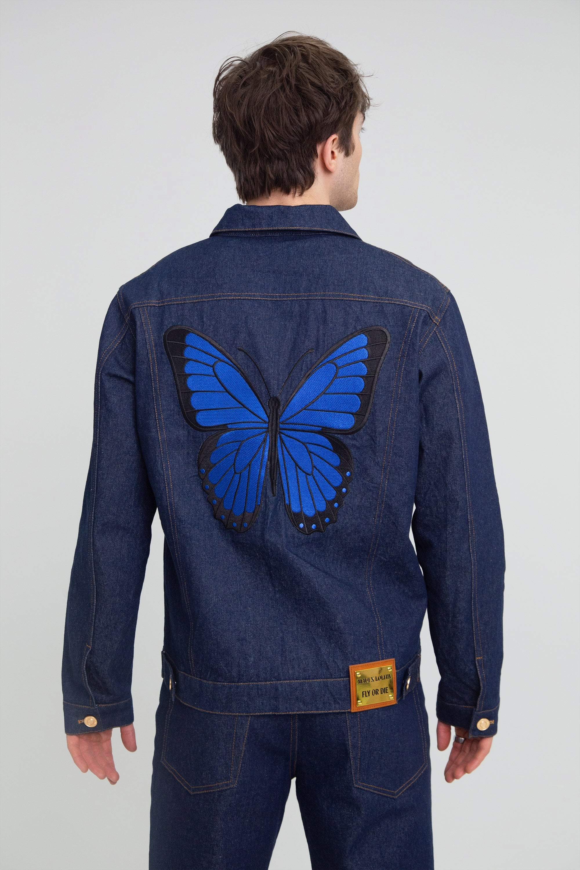 Person wearing a blue denim jacket with a large blue butterfly design on the back against a plain background