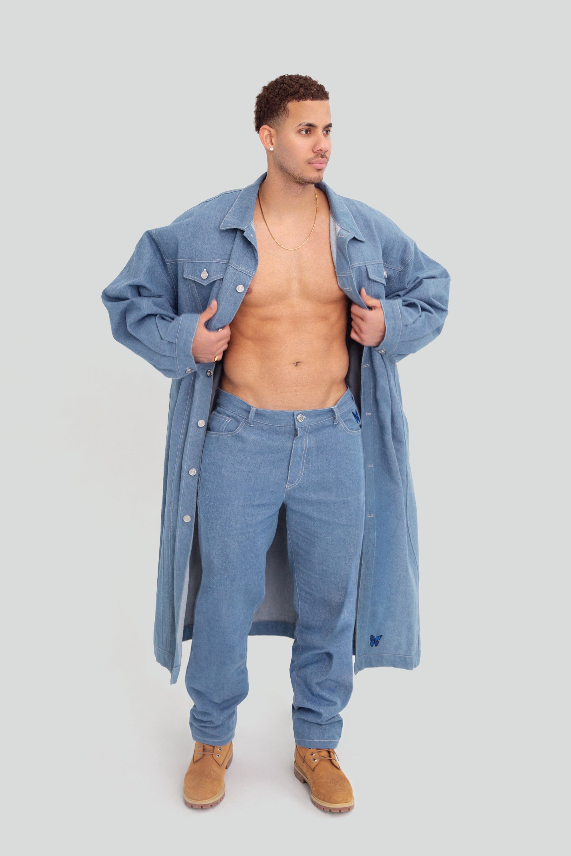 Man wearing a long denim jacket over jeans on a plain background