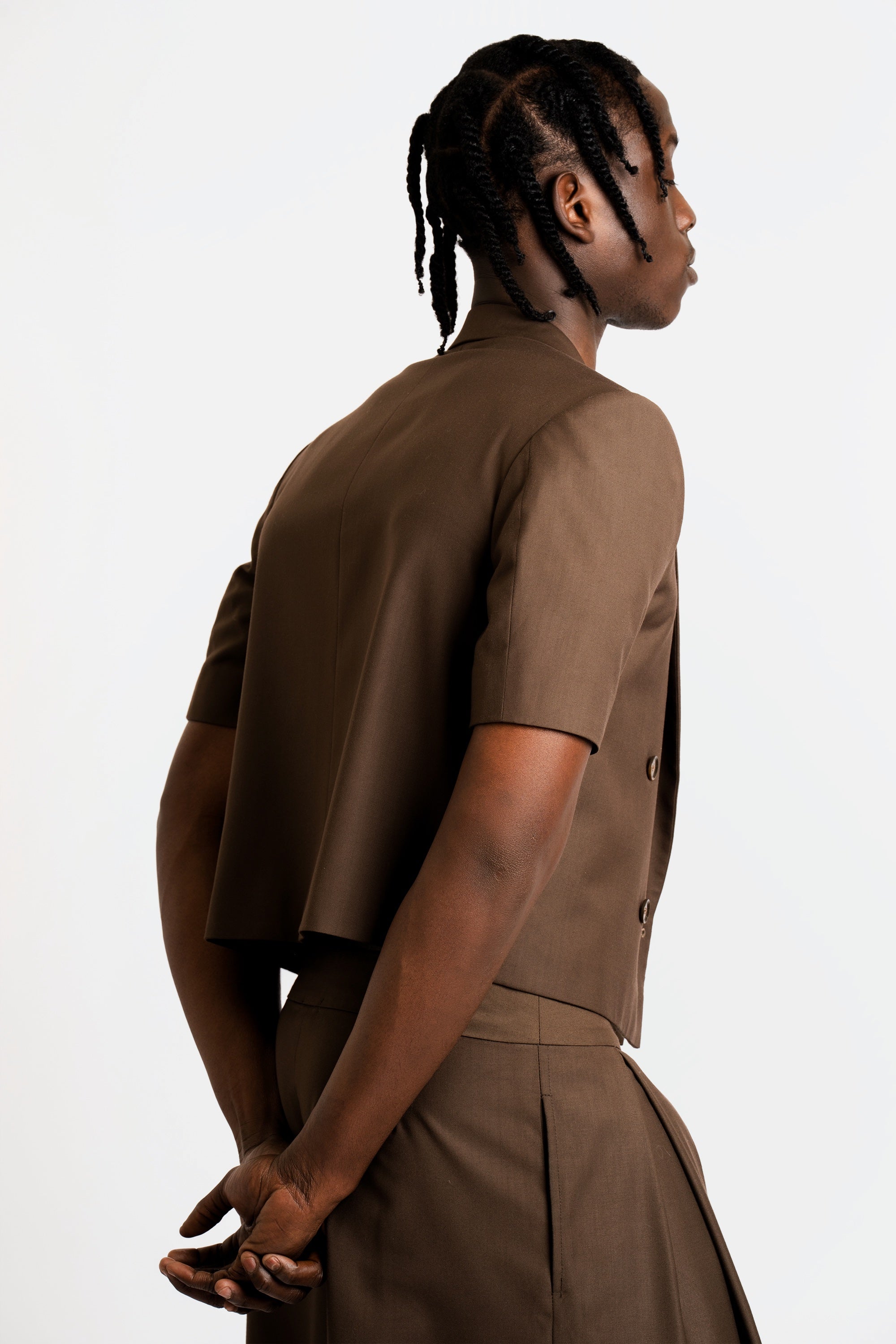 Man wearing a cropped brown blazer and short set on a white background with his back towards the camera.
