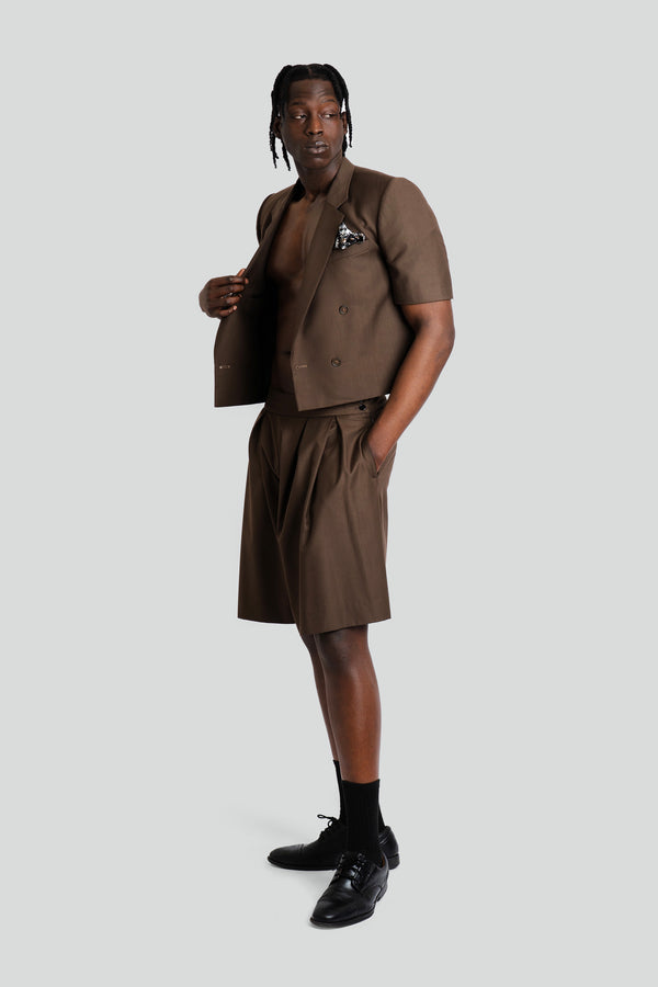 A full-length studio shot of a model wearing a tan cropped suit set featuring a double-breasted blazer with sharp lapels paired with dramatic high-waisted, pleated wide-leg trousers in an earthy taupe hue