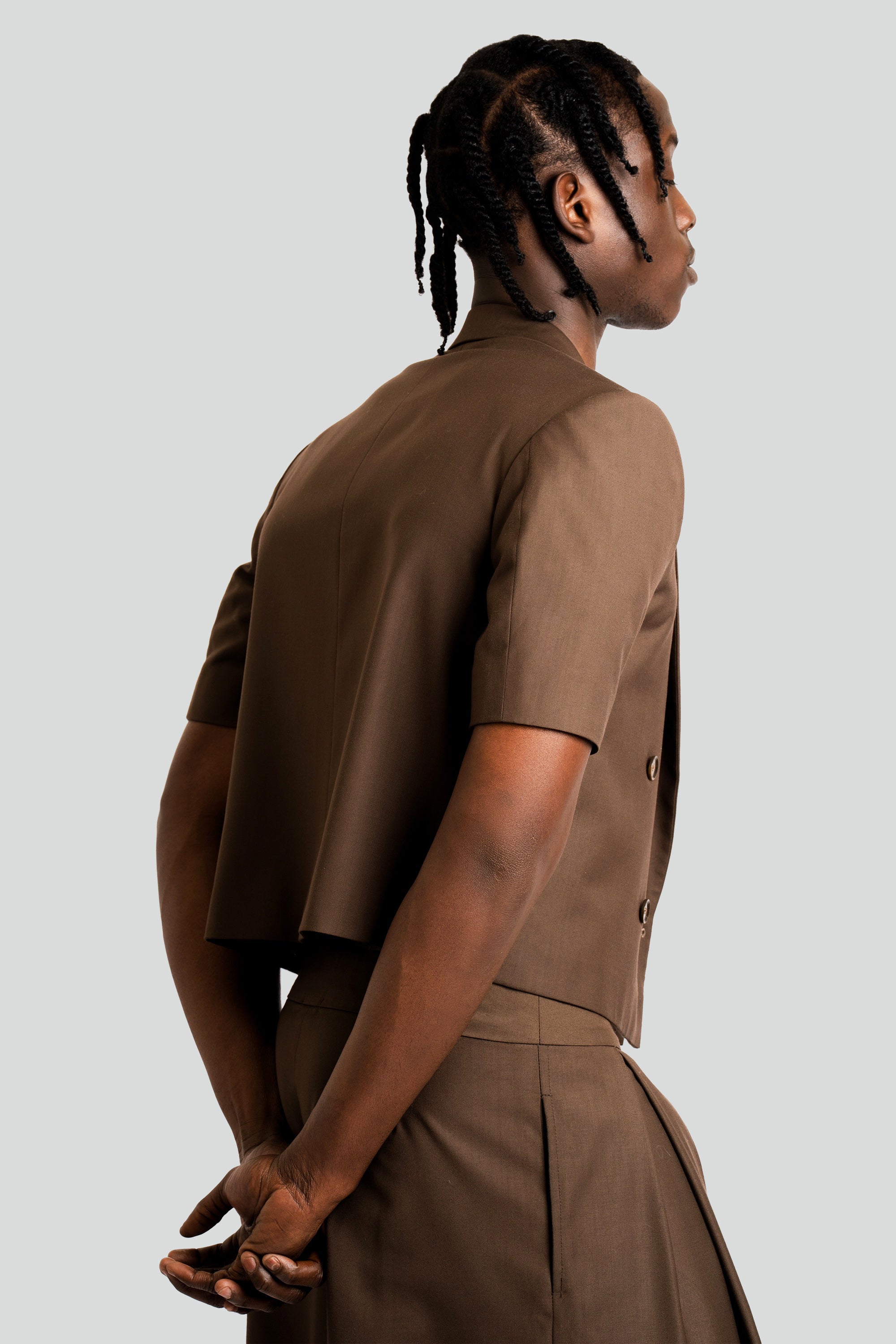 Man wearing a cropped brown blazer and short set on a white background with his back towards the camera.