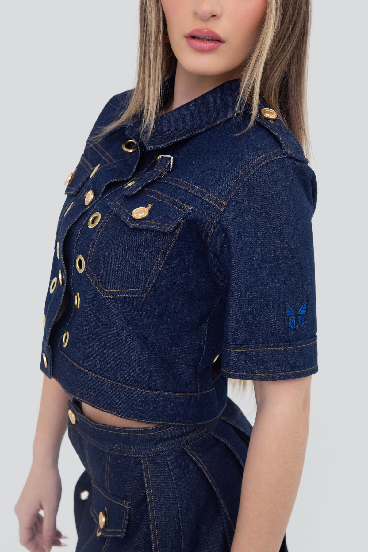 Close up view of a structured dark wash indigo denim cropped shirt highlighting the clean stitching and signature gold grommet details