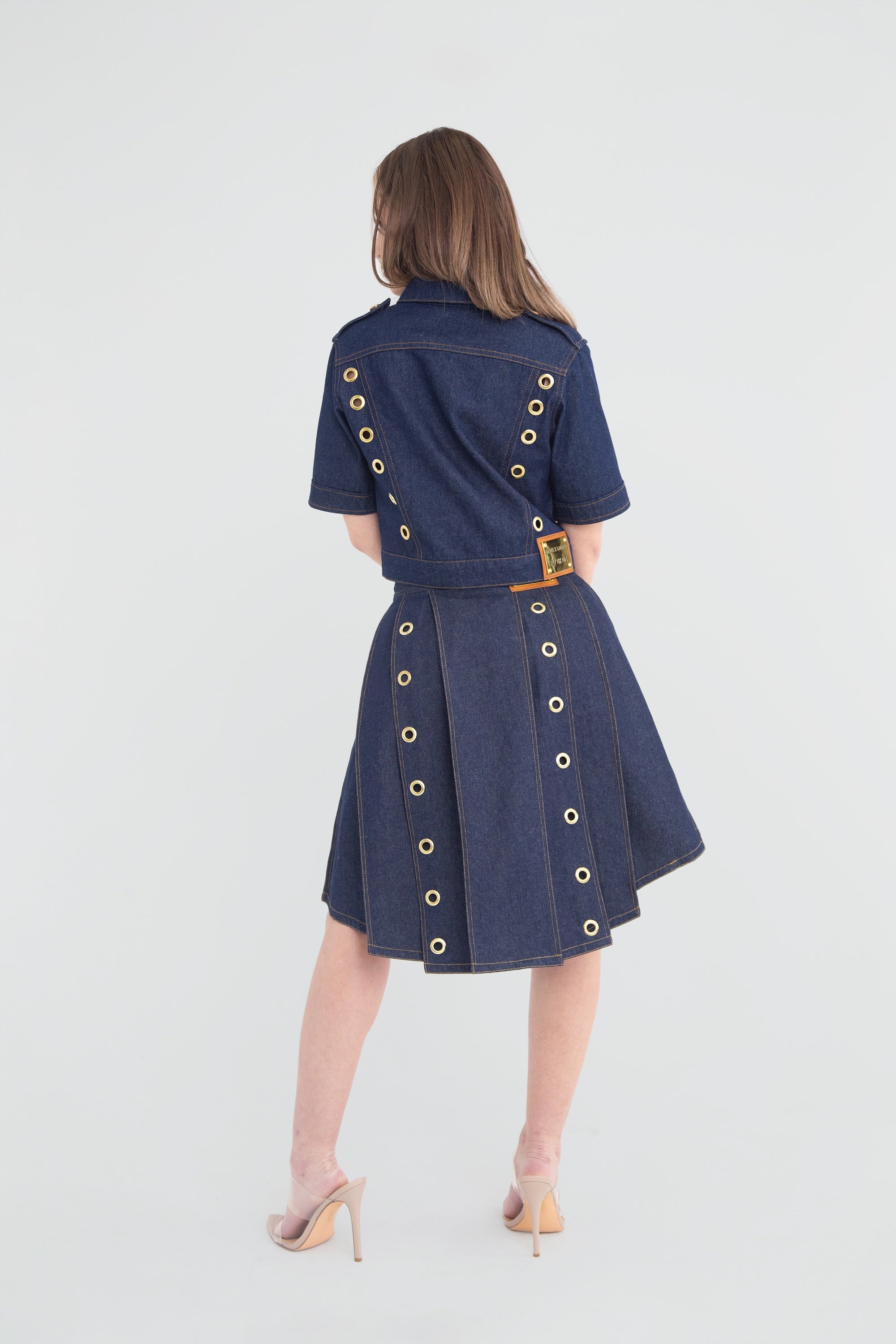 Woman wearing a denim outfit with gold grommets on a plain background