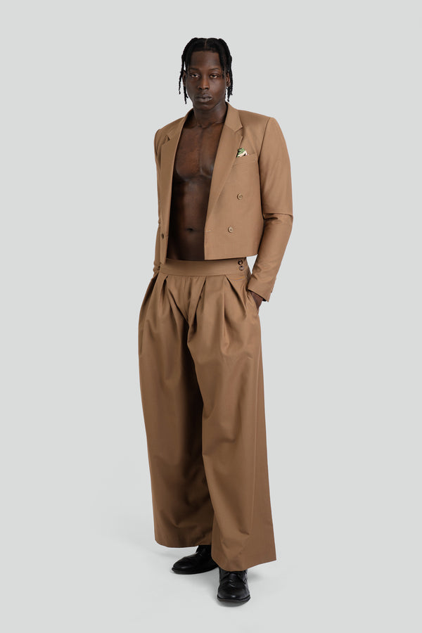 A full-length studio shot of a model wearing a tan cropped suit set featuring a double-breasted blazer with sharp lapels paired with dramatic high-waisted, pleated wide-leg trousers in an earthy taupe hue