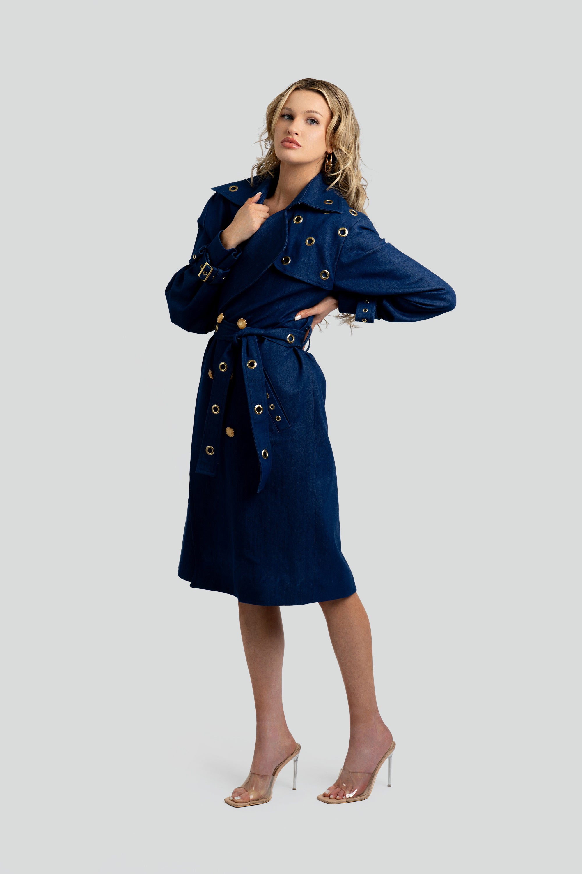 Woman wearing a denim trench coat with a belt and gold accents on a white background