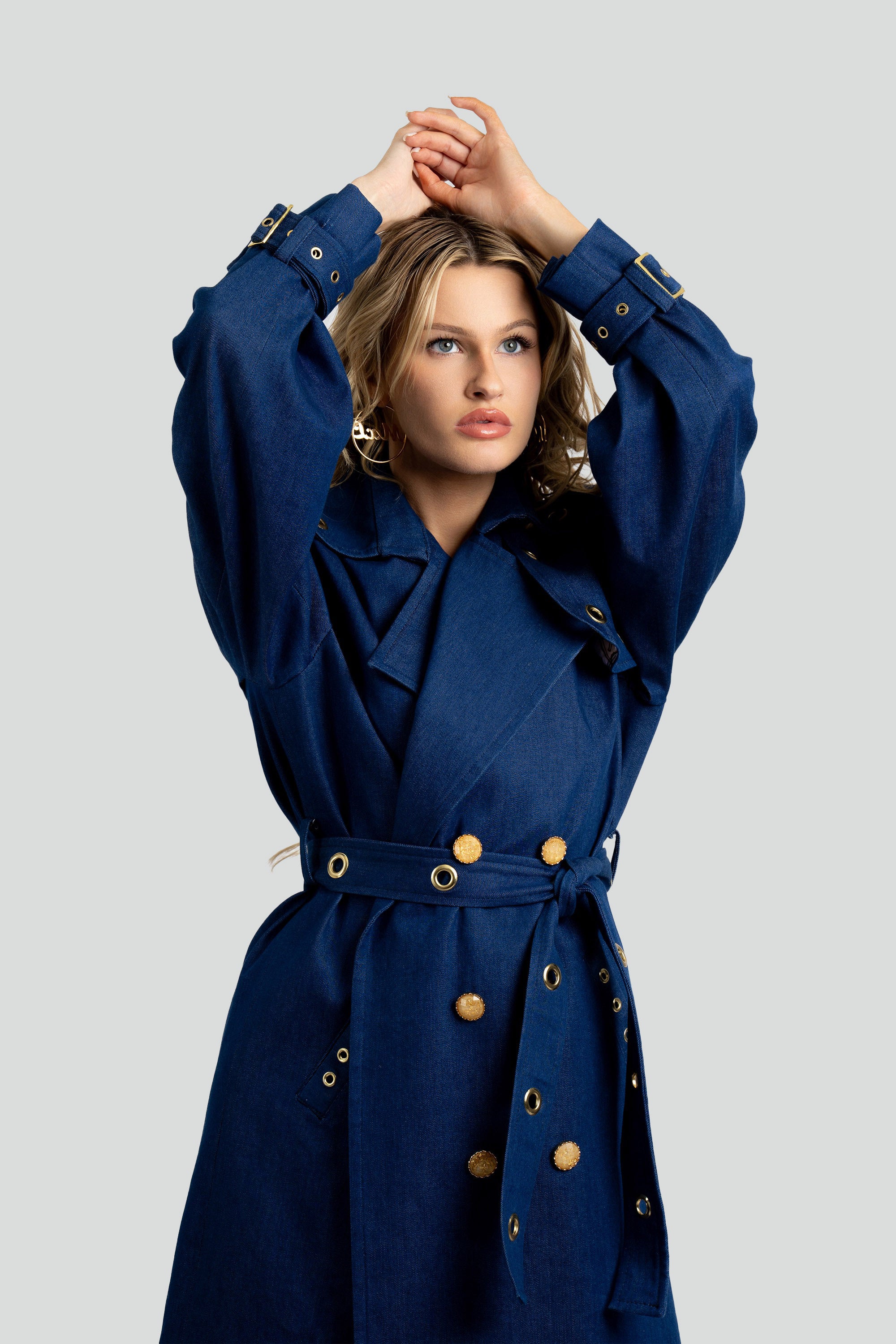 Woman wearing a denim trench coat with a belt and gold accents on a white background