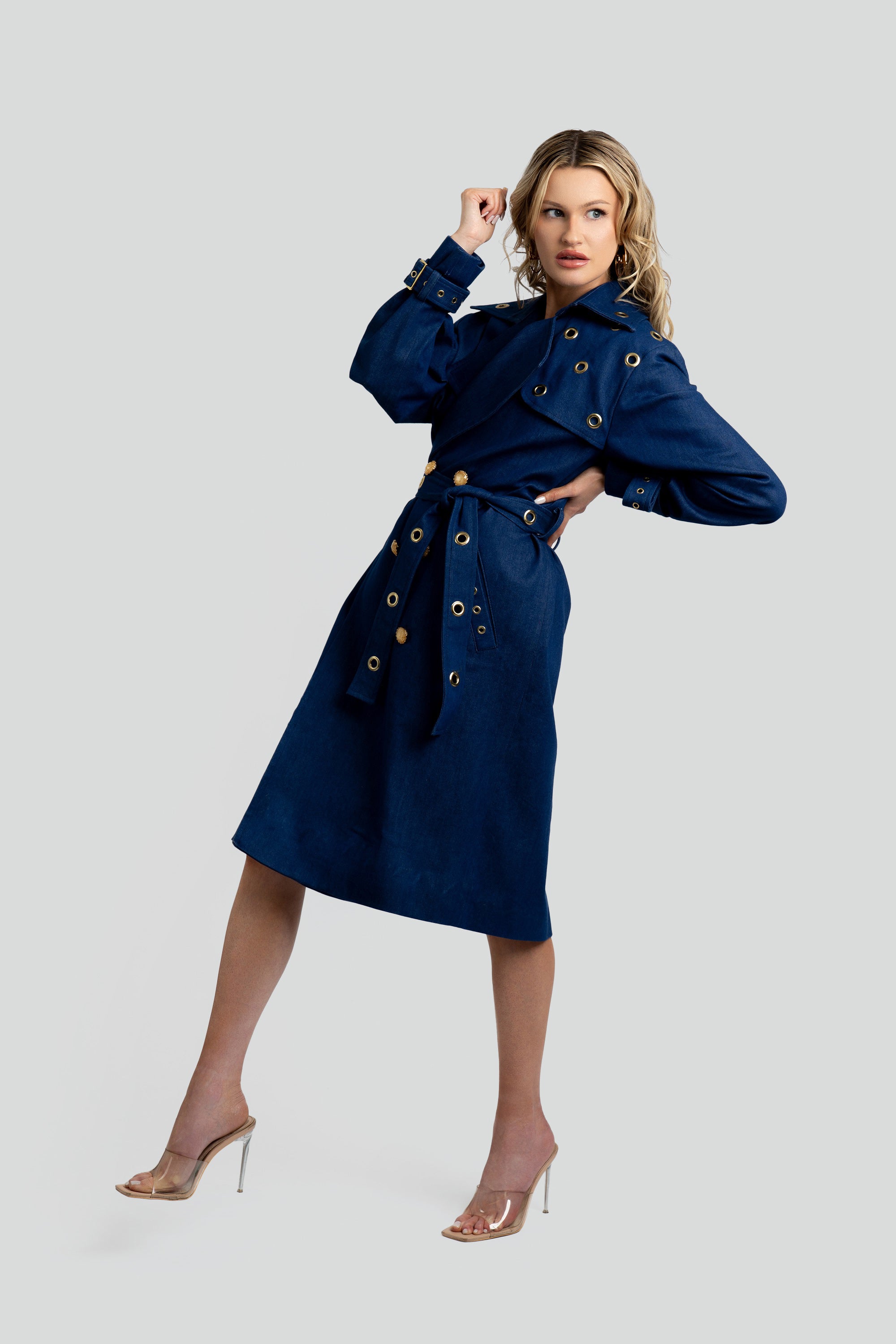 Woman wearing a denim trench coat with a belt and gold accents on a white background