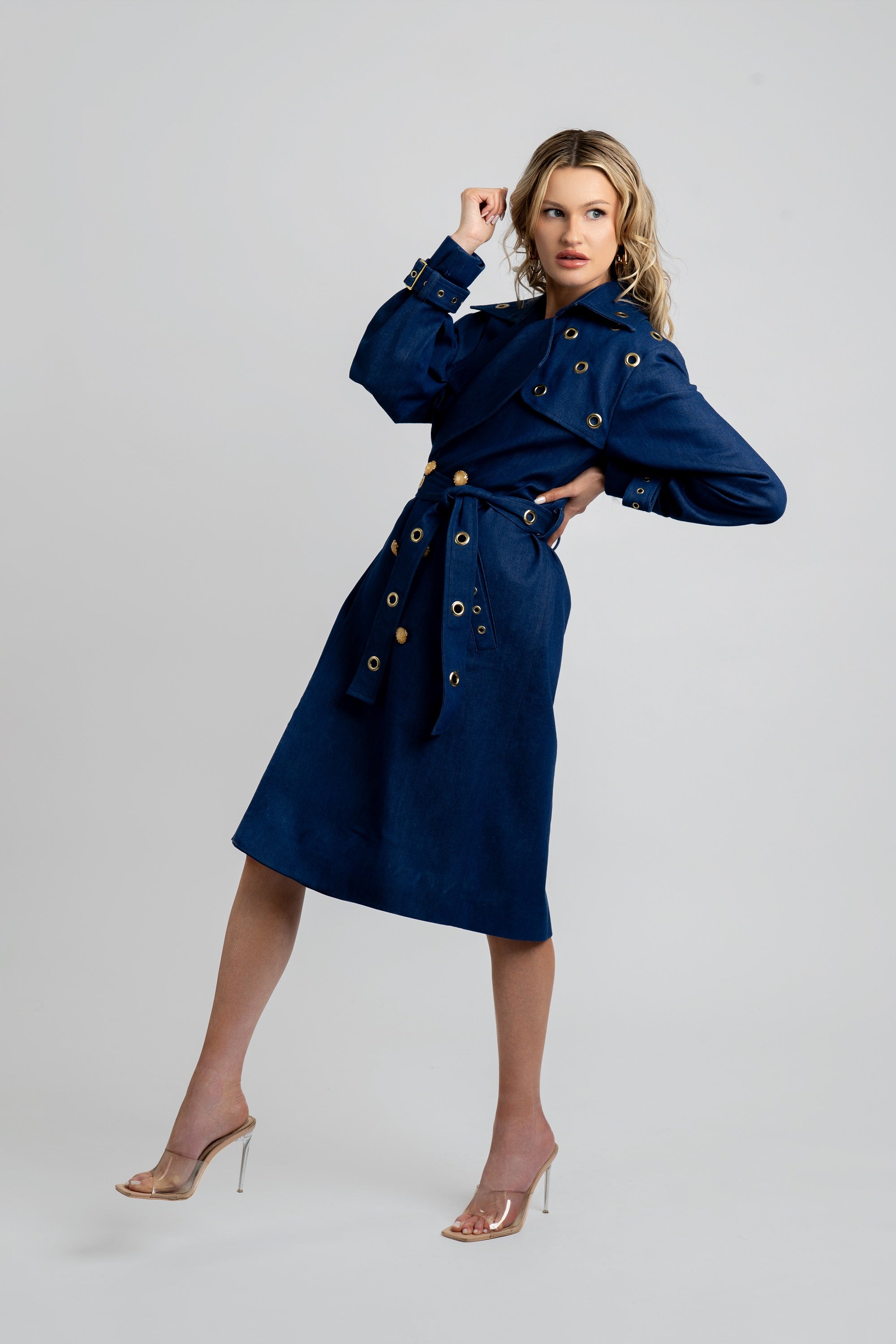 Woman wearing a denim trench coat with a belt and gold accents on a white background