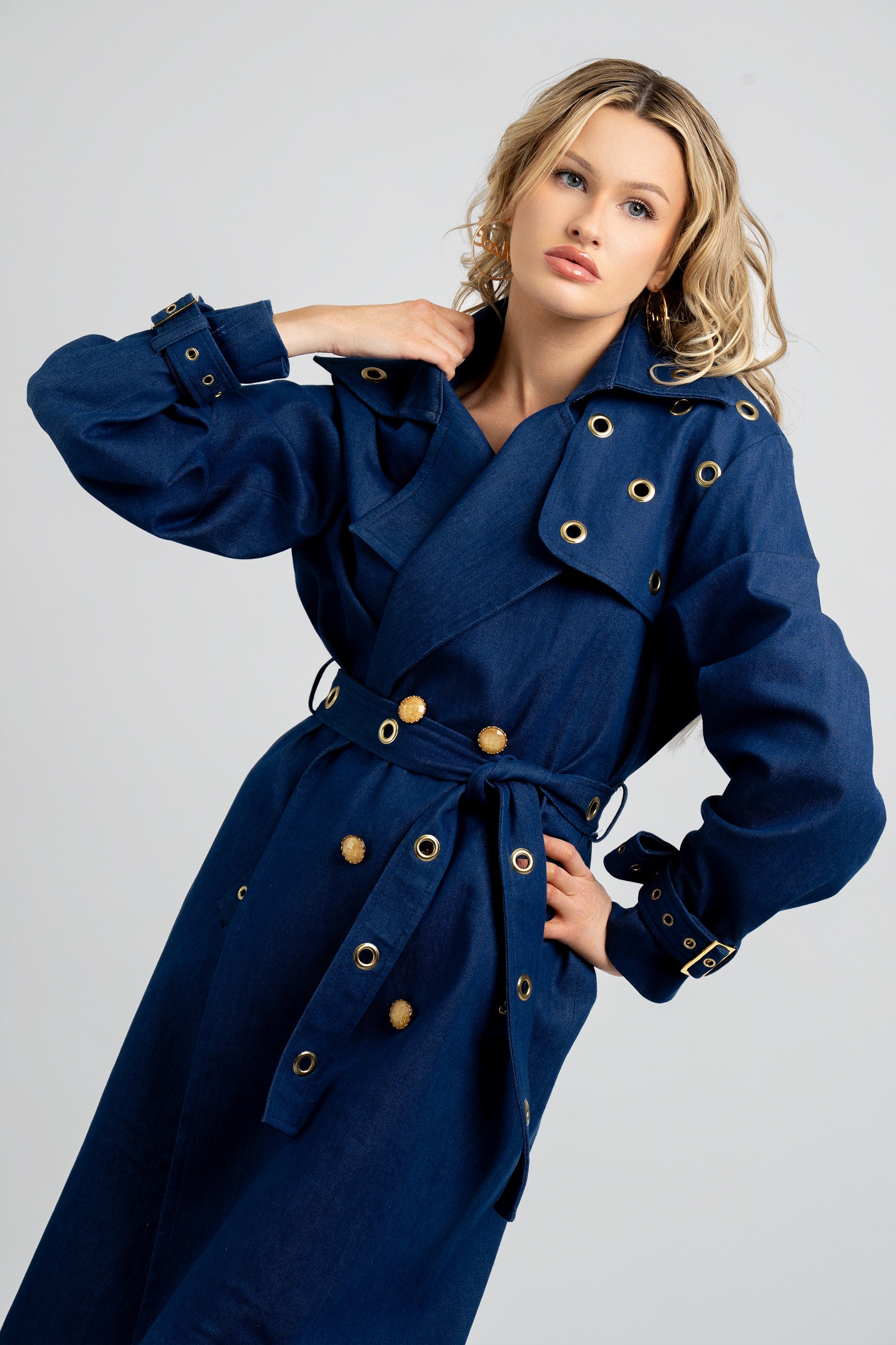 Woman wearing a denim trench coat with a belt and gold accents on a white background
