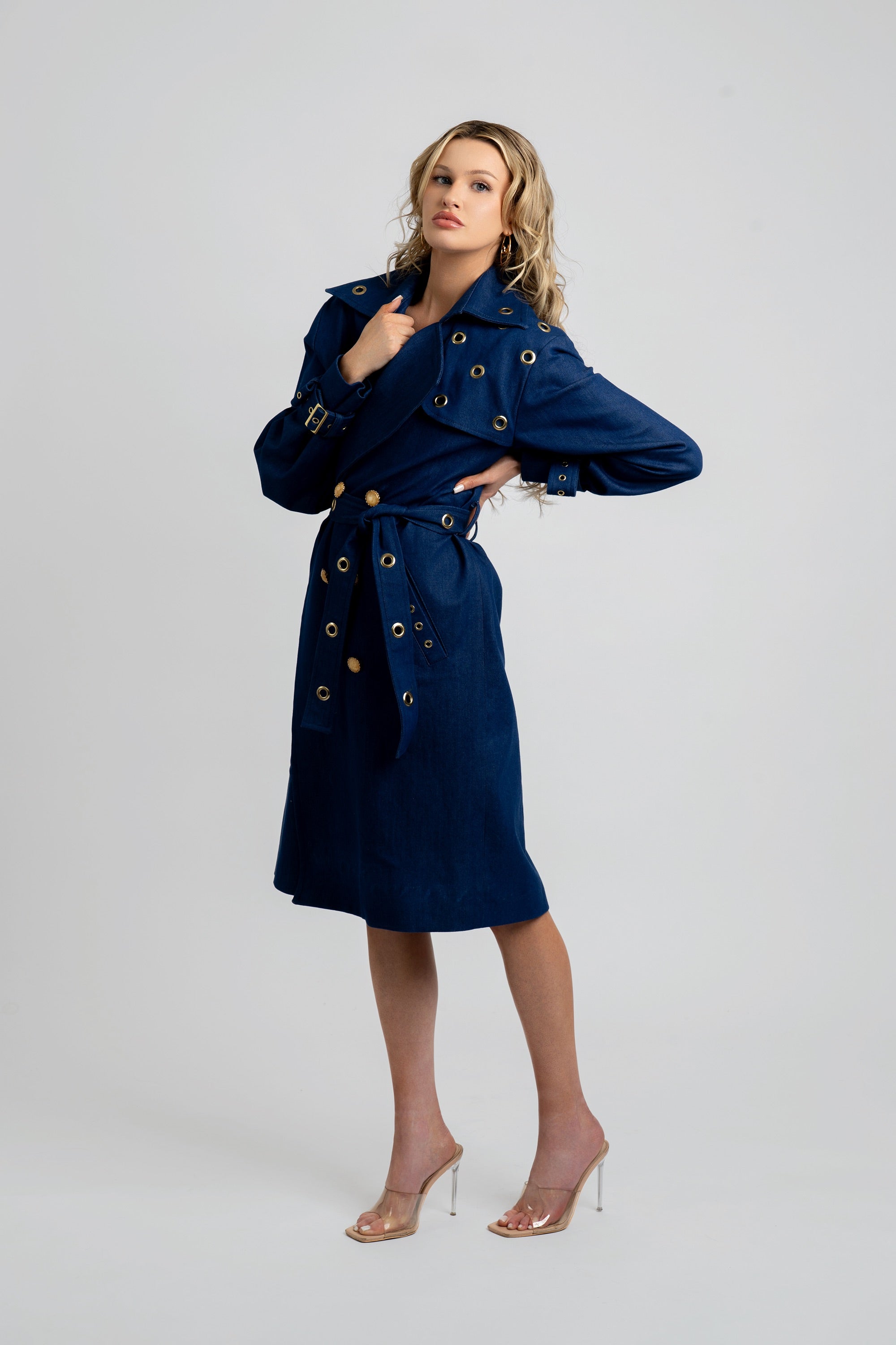 Woman wearing a denim trench coat with a belt and gold accents on a white background