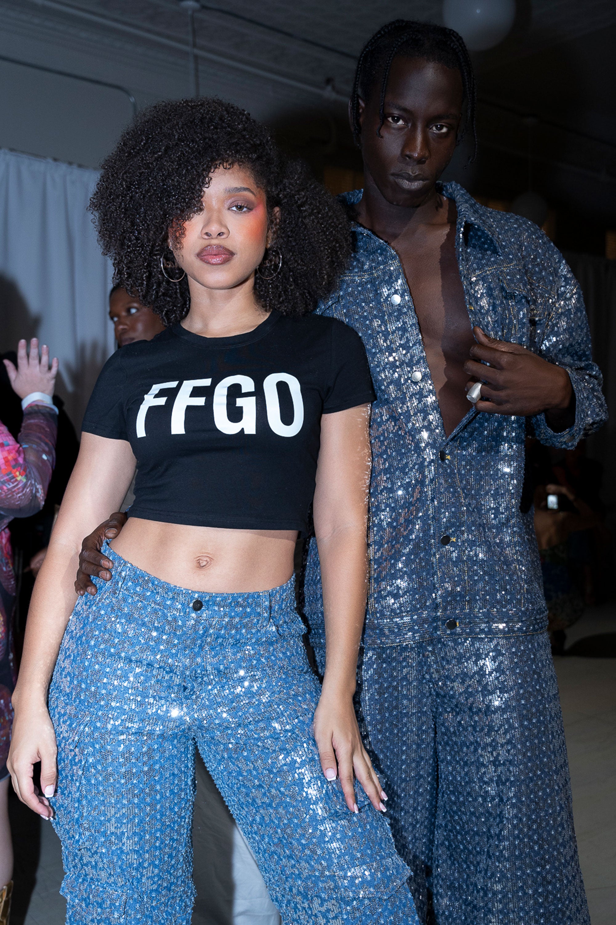 Two models wearing blue sequined denim clothes. The woman on the left is wearing a black top with 'FFGO' text and the man on the right is wearing a full denim set.
