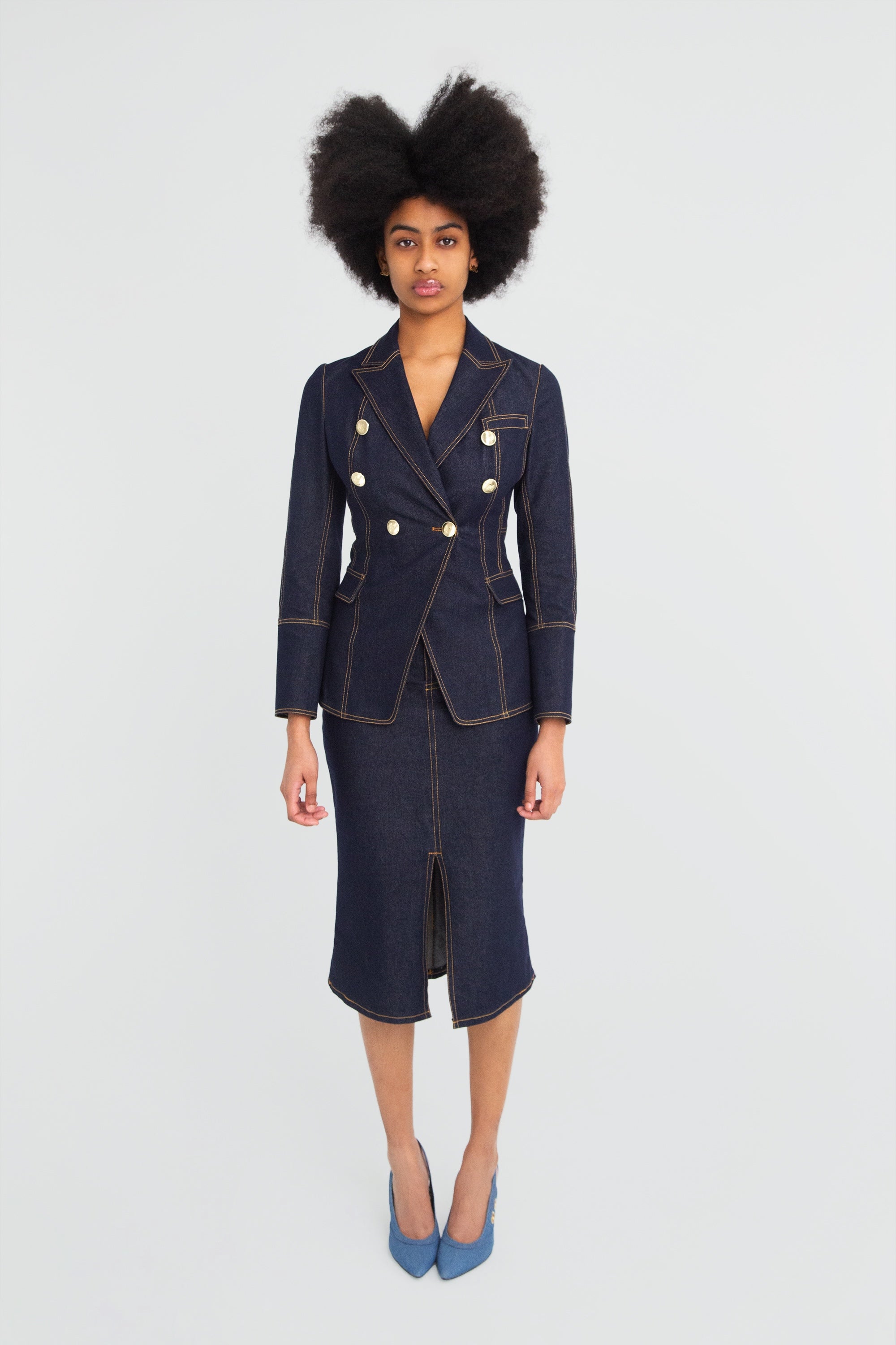 Woman wearing a dark denim blazer with gold buttons and pencil skirt on a plain background