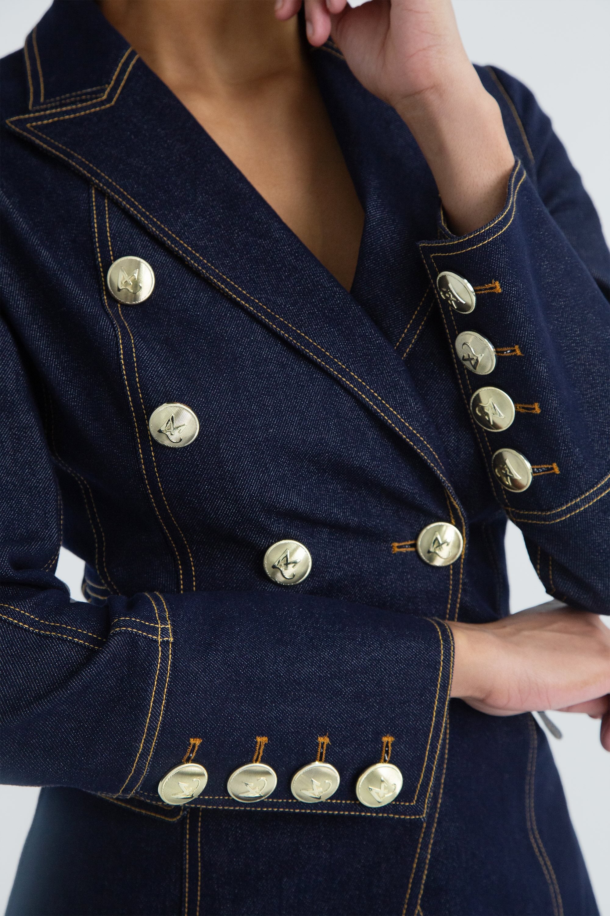 Close up of a dark denim blazer with gold buttons worn by a model on a plain background