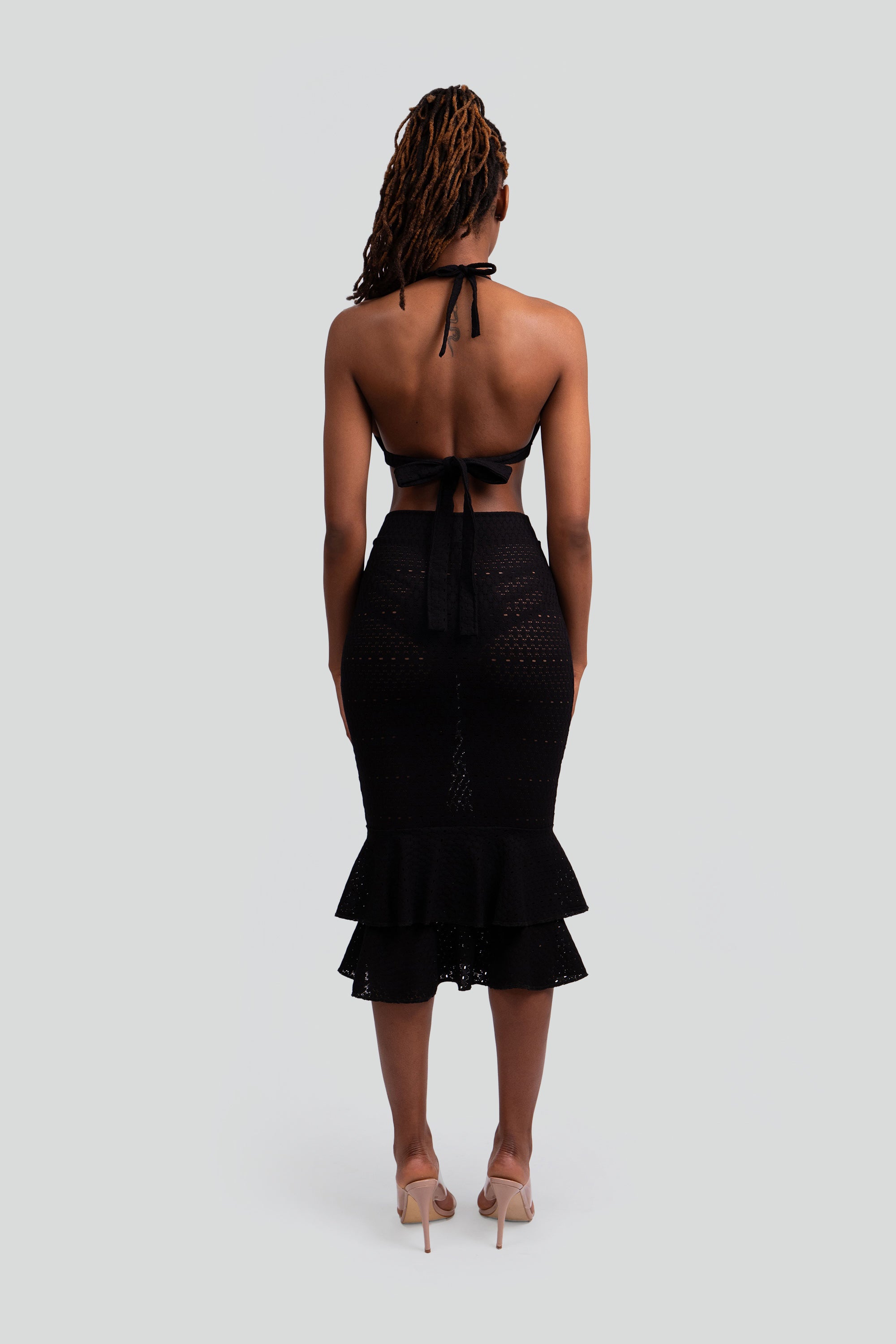 Back view of a woman wearing a black lace knit outfit with a halter top and ruffled skirt on a white background