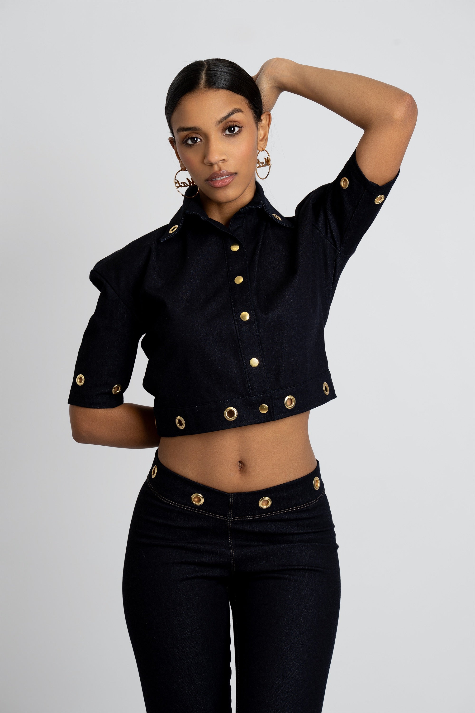 Woman wearing a dark denim outfit with gold embellishments on a plain background