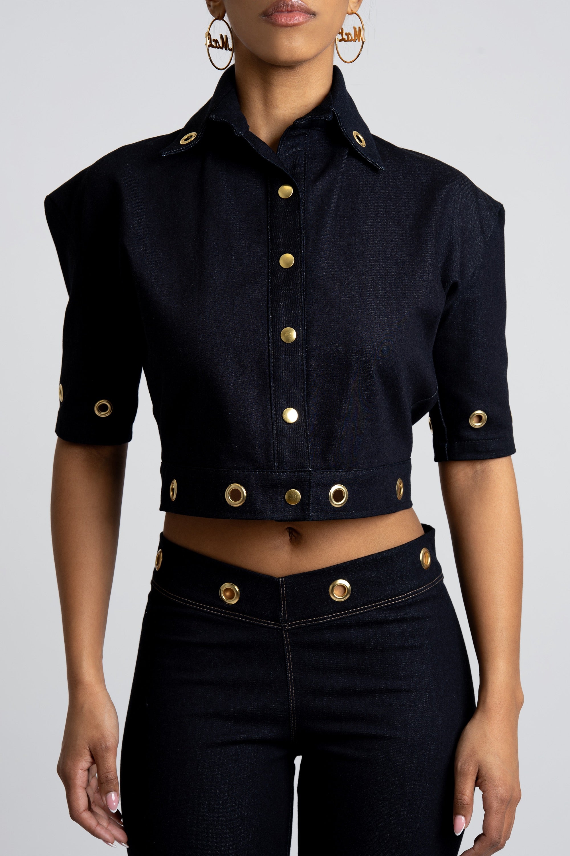 Cropped dark denim shirt with gold embellishments worn by a person on a gray background