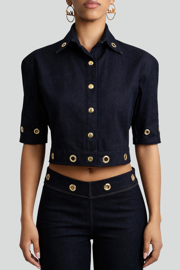 Woman wearing a dark wash indigo denim cropped shirt featuring structured angular shoulders and metallic grommet detailing
