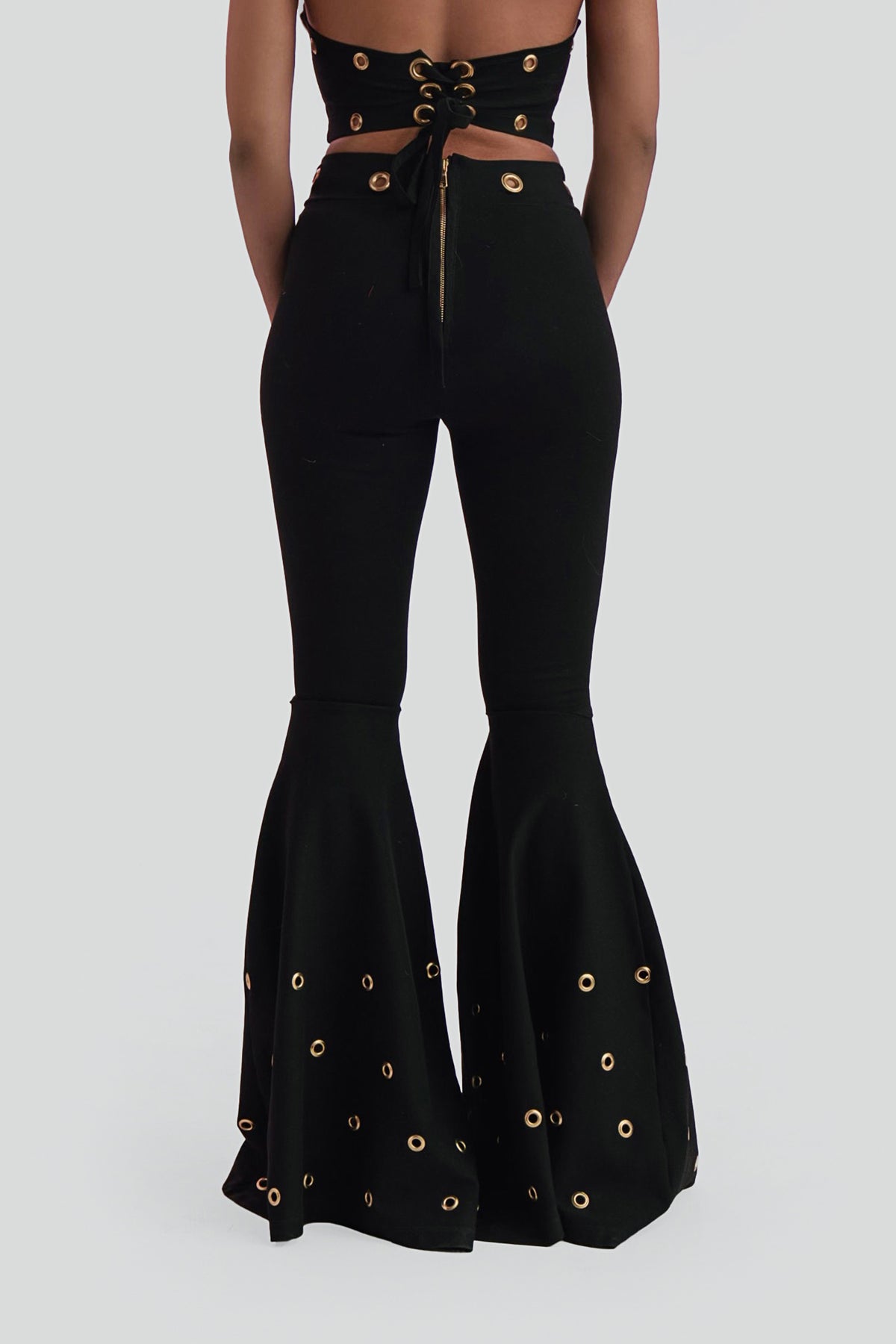 High-waisted black bell-bottom pants with a dramatic flared leg and gold grommet accents styled for an editorial look