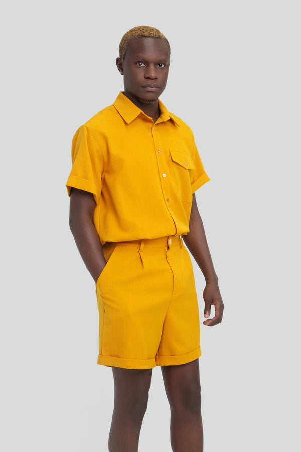 Man wearing a mustard yellow short suit set featuring a relaxed short sleeve button up shirt and matching high waisted pleated shorts