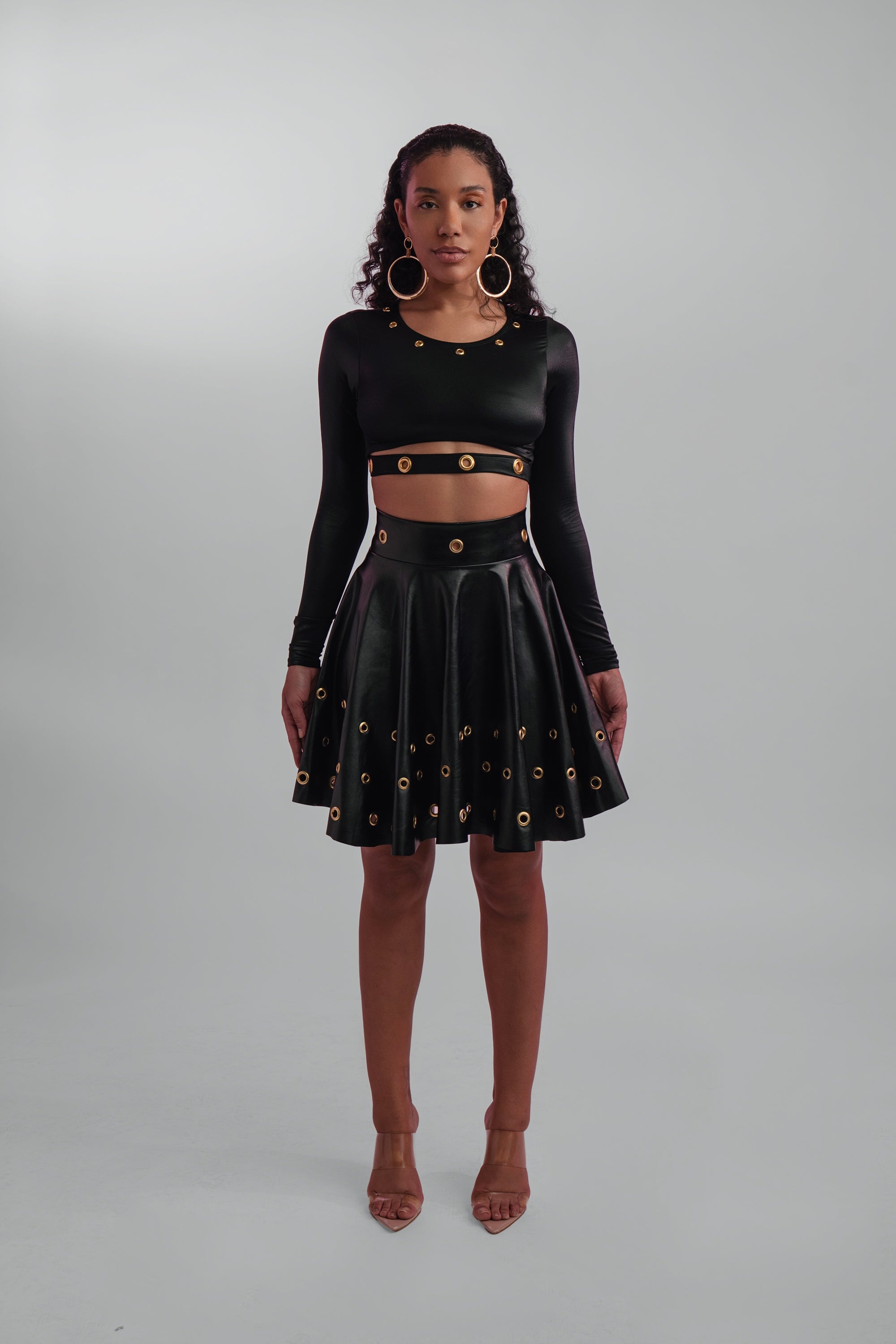 Woman wearing a black long-sleeve top and black skirt with gold grommets on a gray background
