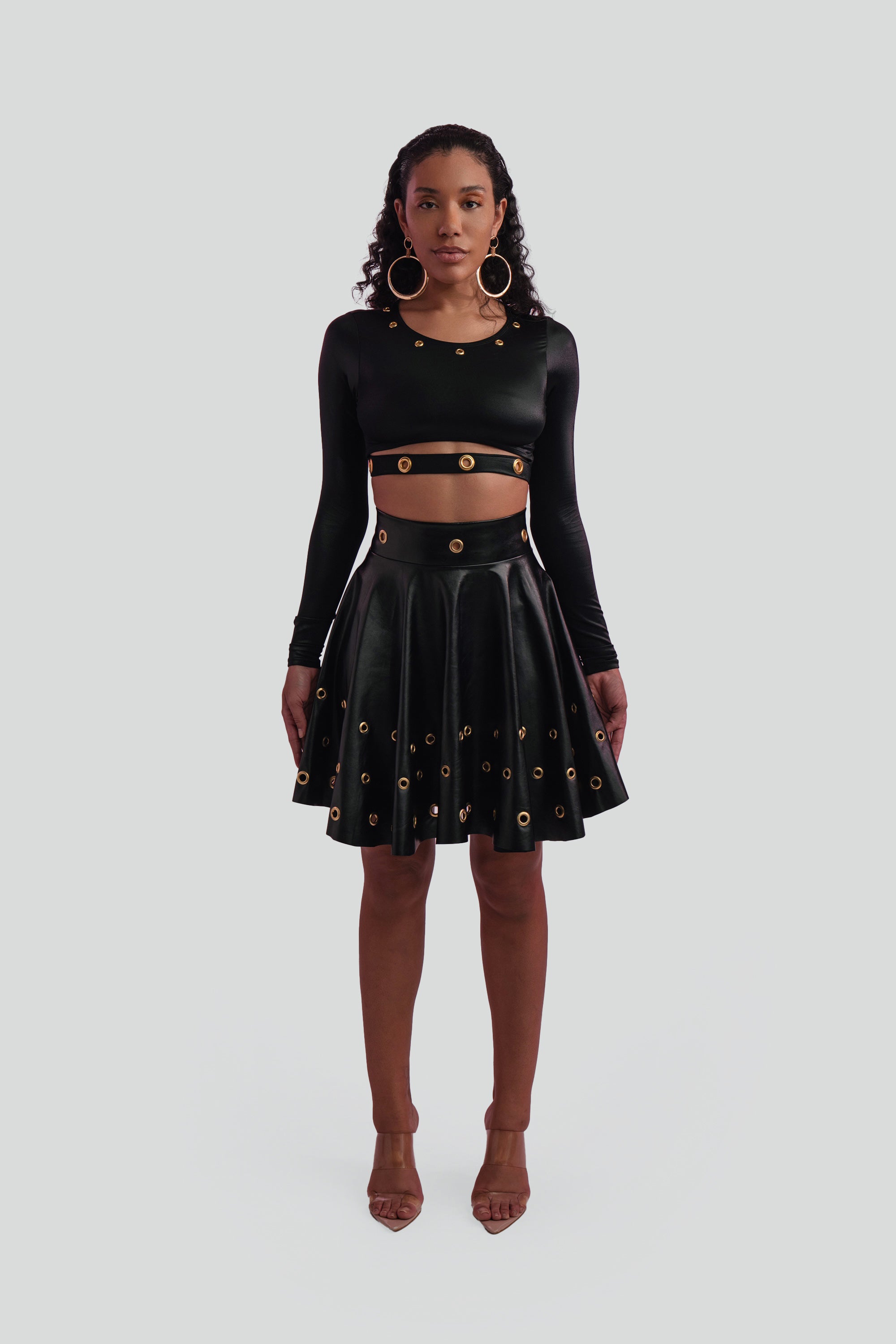 Woman wearing a black long-sleeve top and black skirt with gold grommets on a gray background