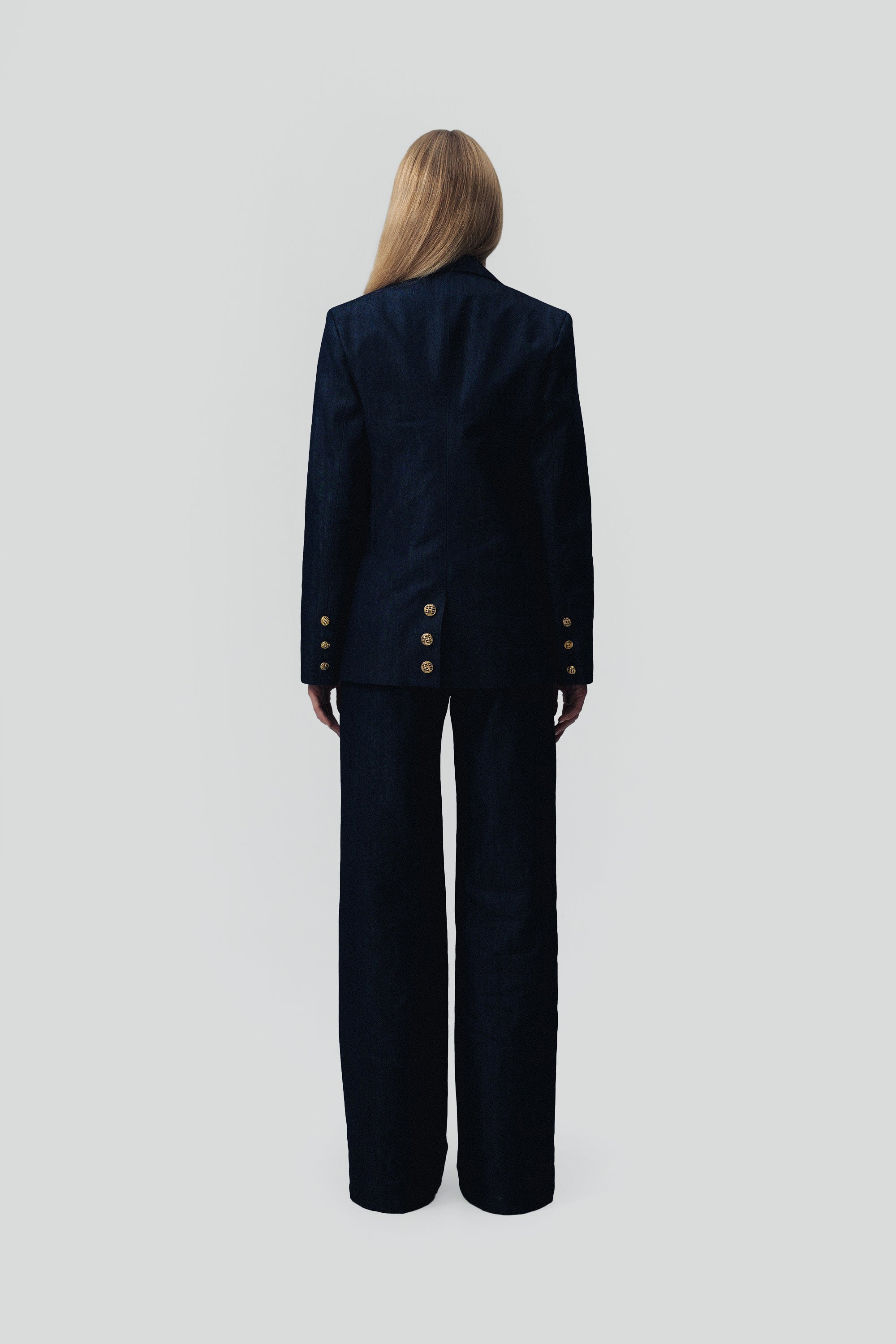 Back view of a woman wearing a dark blue denim suit with gold buttons on a gray background