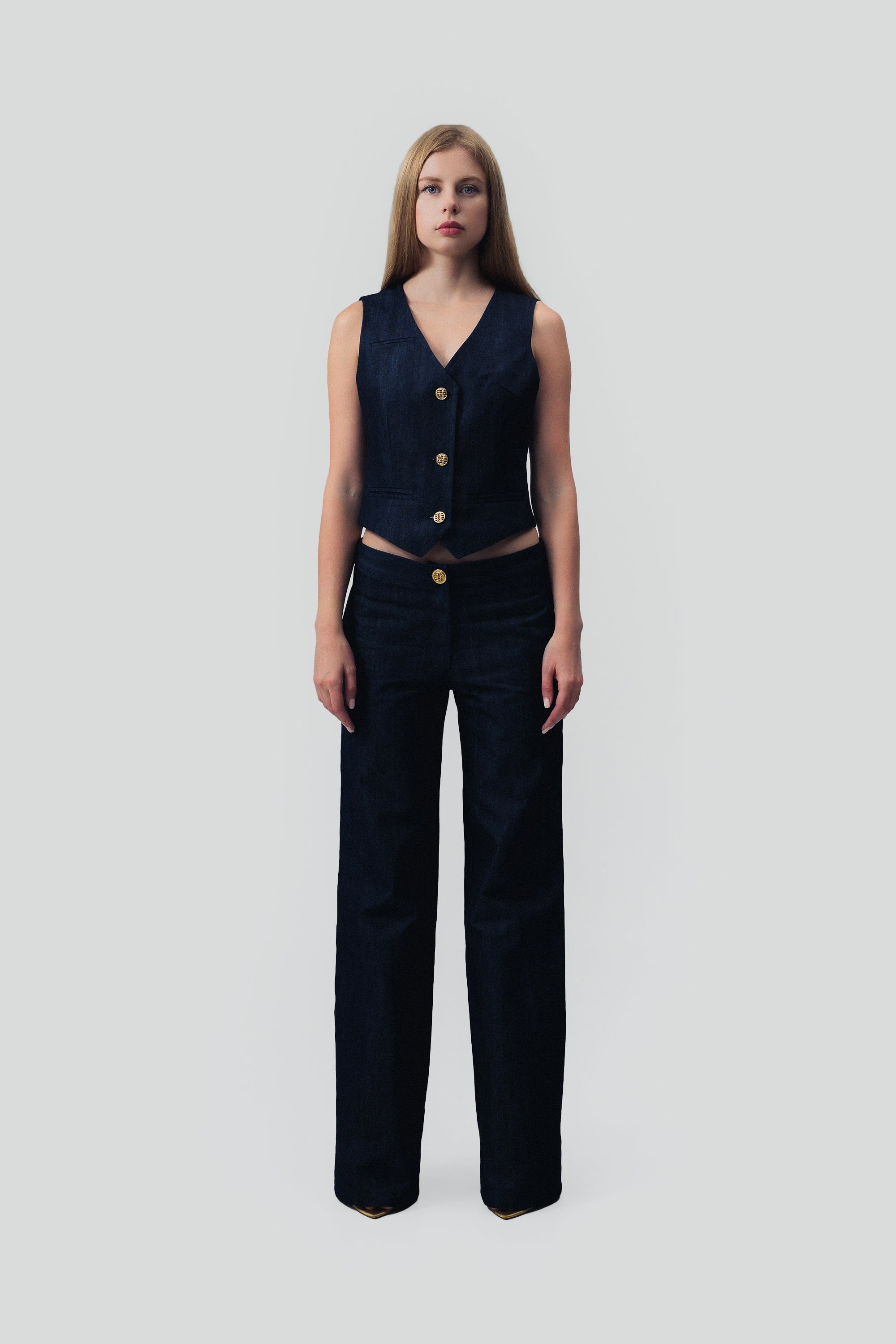 Woman wearing a dark blue denim vest and pants with gold buttons on a grey background