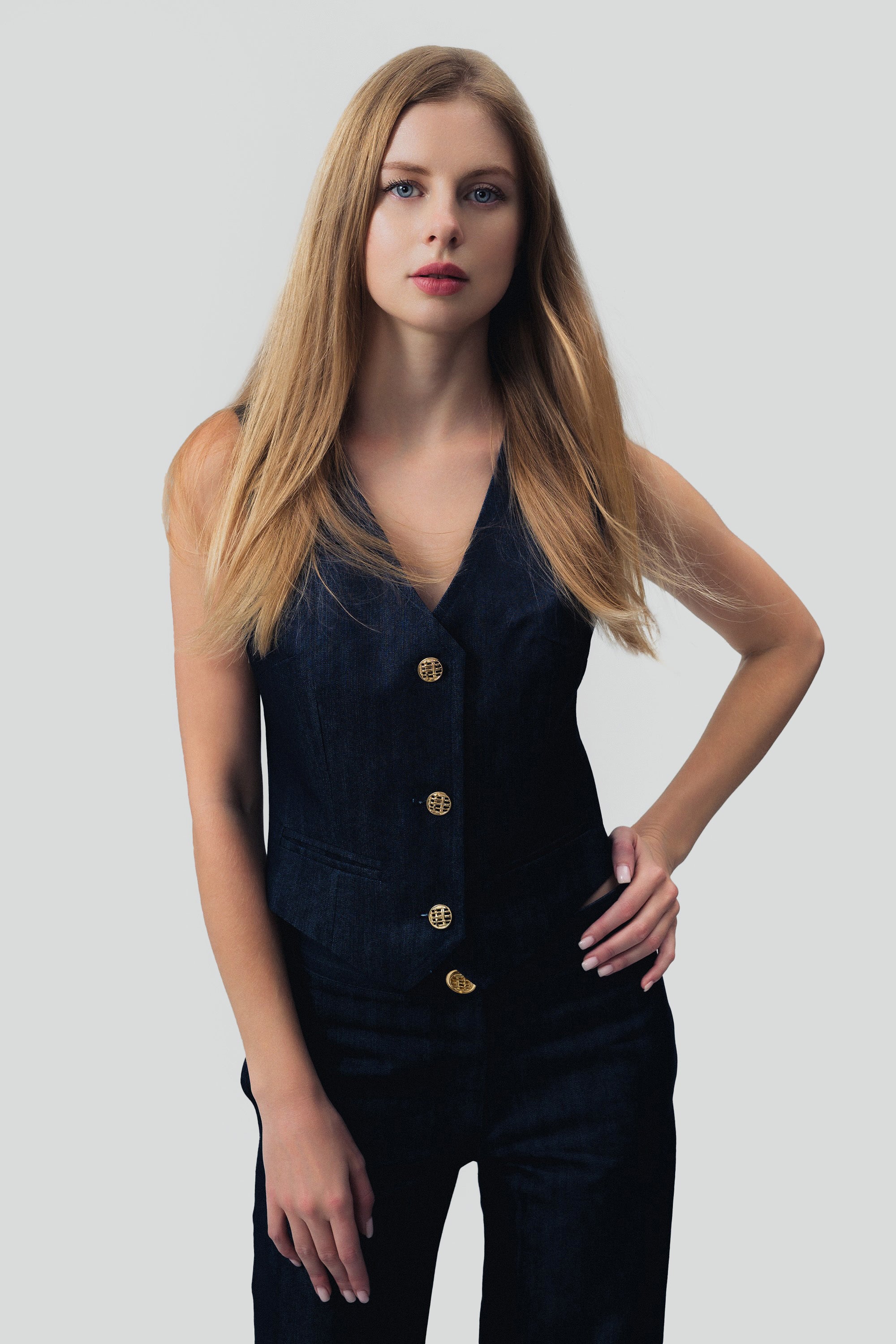 Woman wearing a dark blue denim vest and pants with gold buttons on a grey background