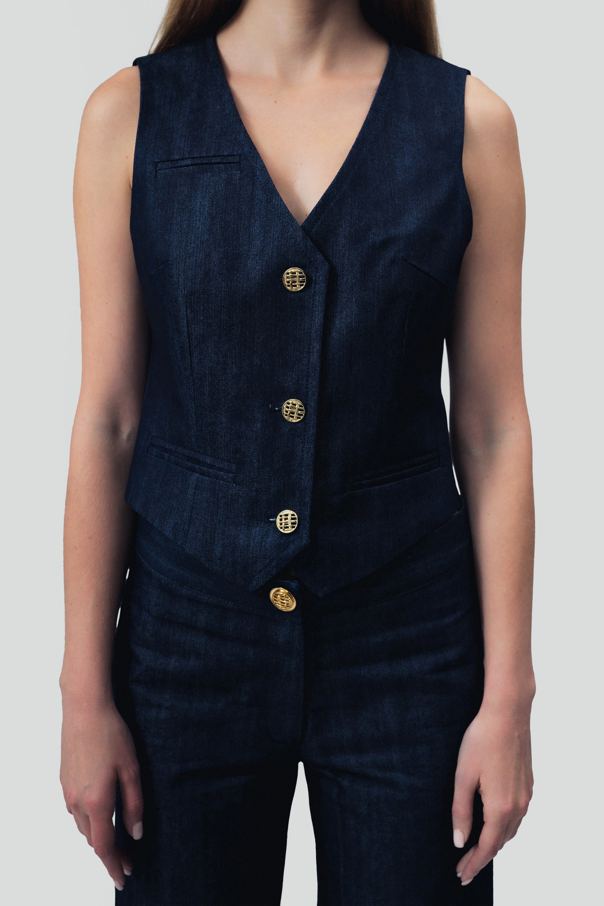 Close up view of a woman wearing a dark blue denim vest and pants with gold buttons on a grey background