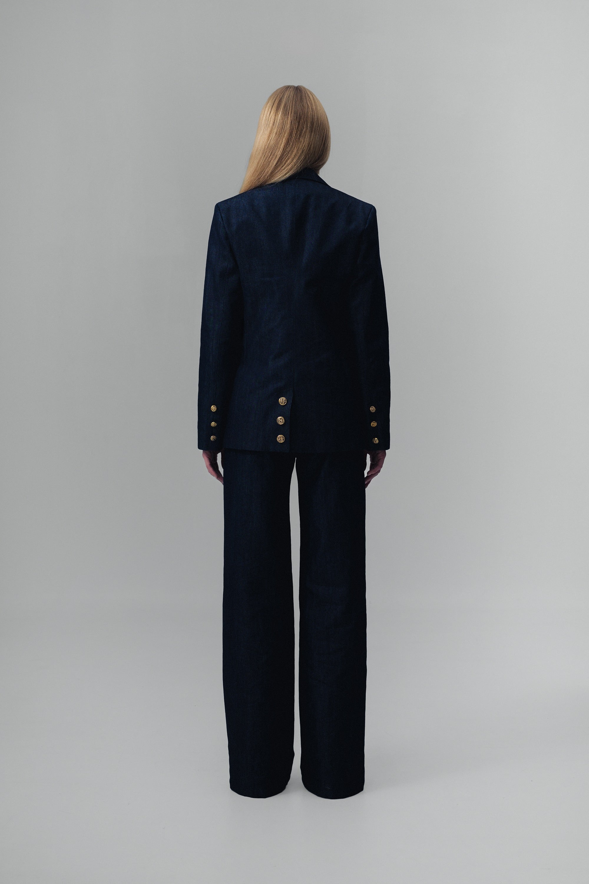 Back view of a woman wearing a dark blue denim suit with gold buttons on a gray background