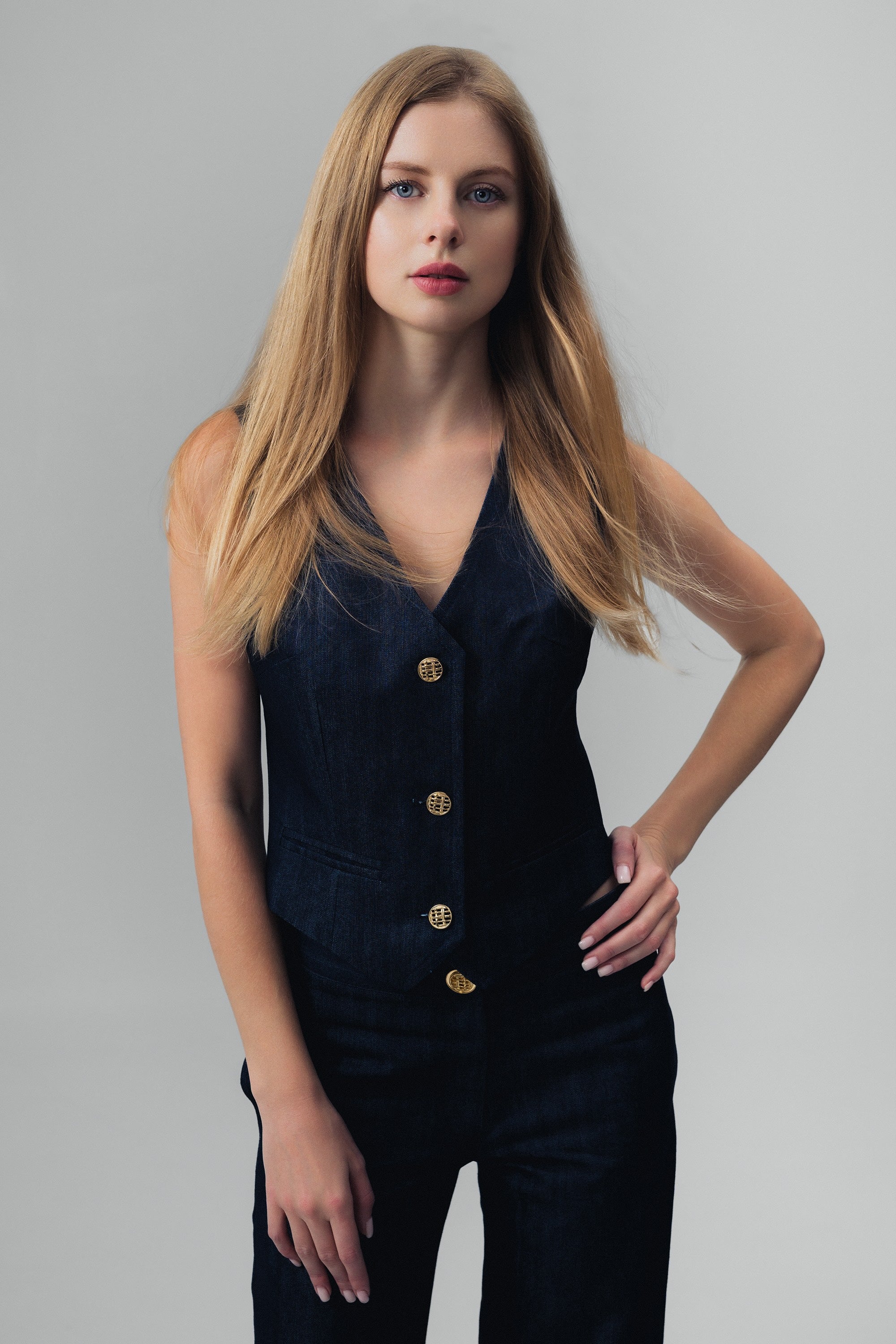 Woman wearing a dark blue denim vest and pants with gold buttons on a grey background