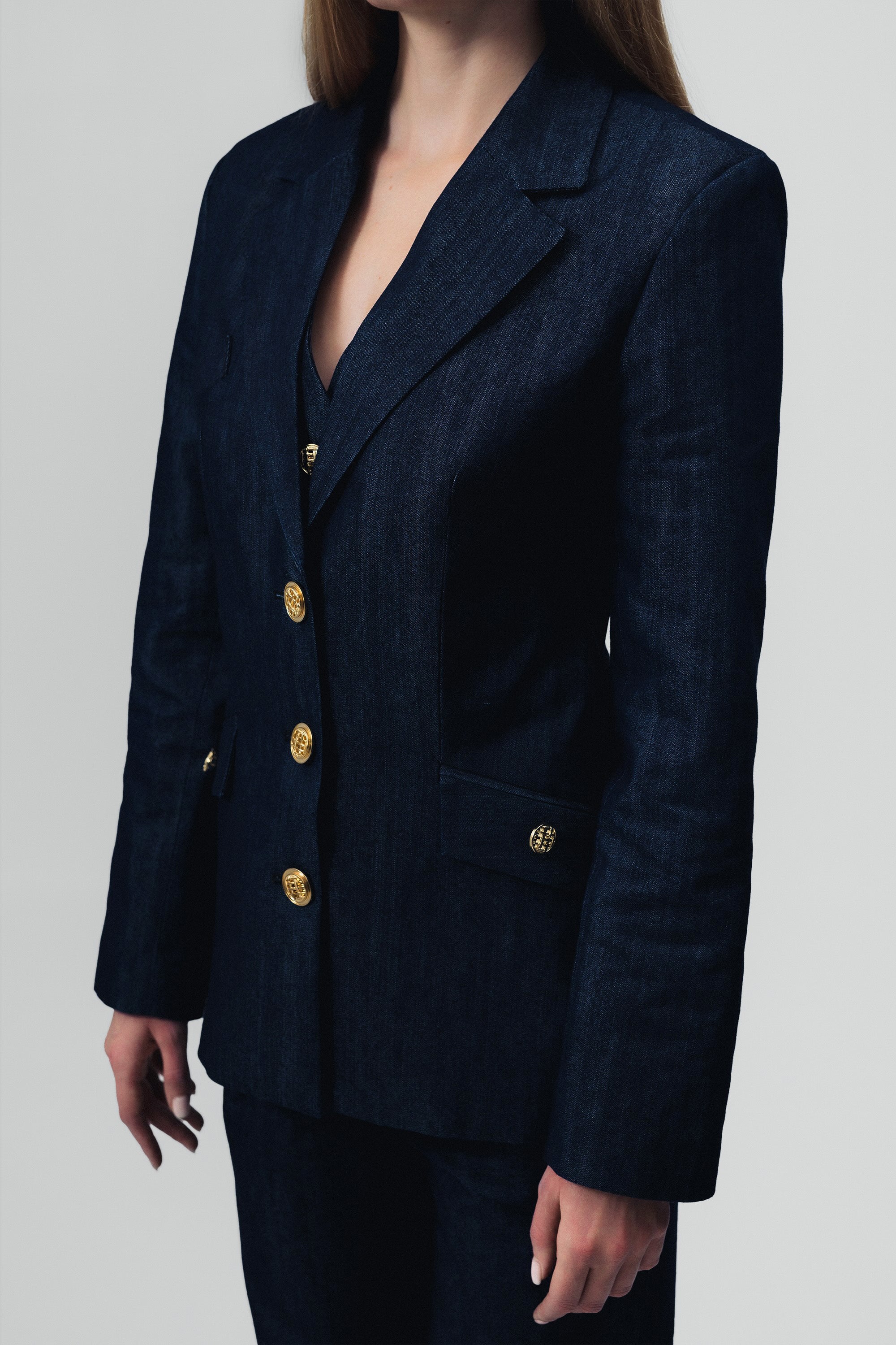 Close up of a woman wearing a dark blue denim blazer with gold buttons on a gray background