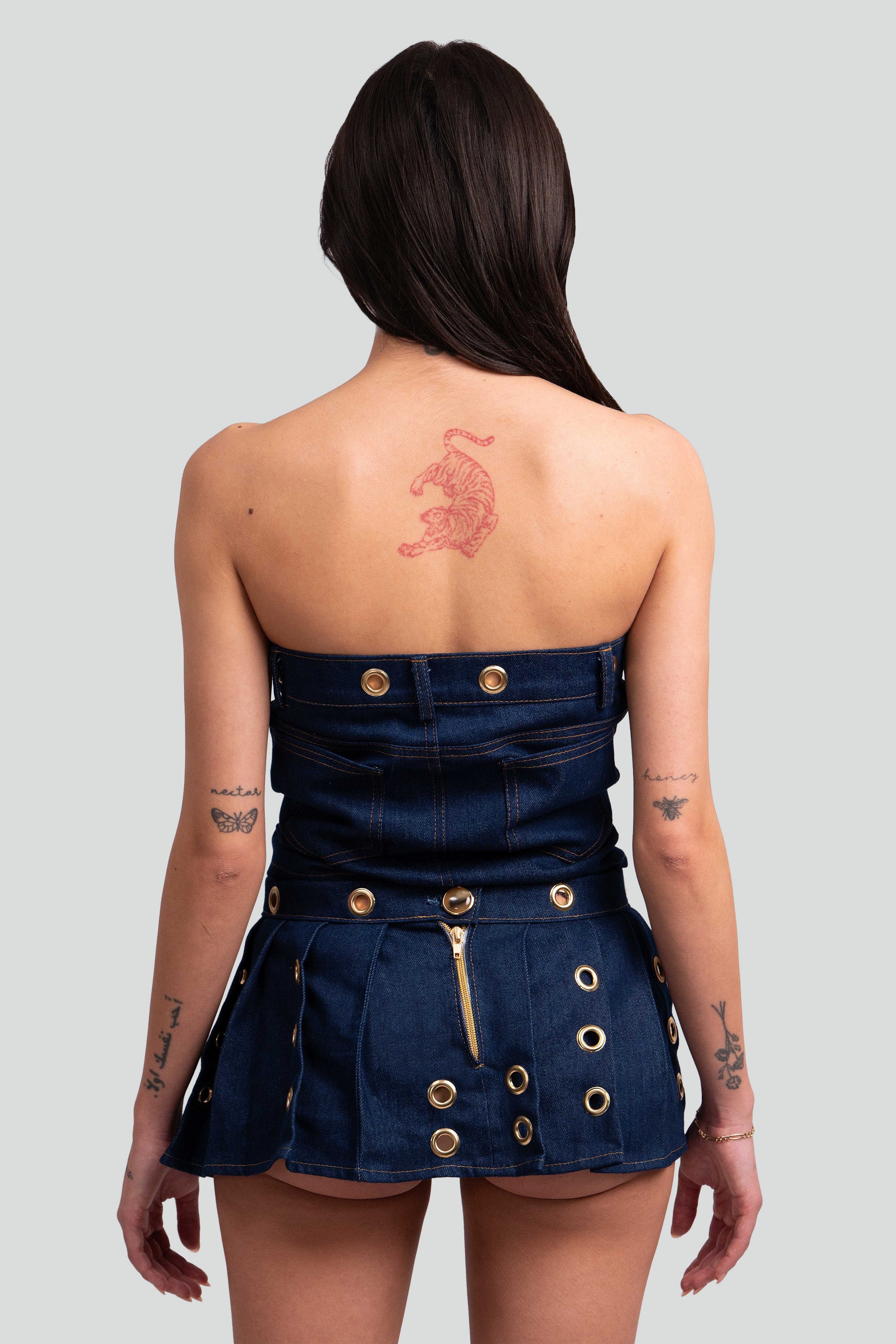 Back view of a woman wearing a denim top and pleated skirt with gold grommets throughout on a white background