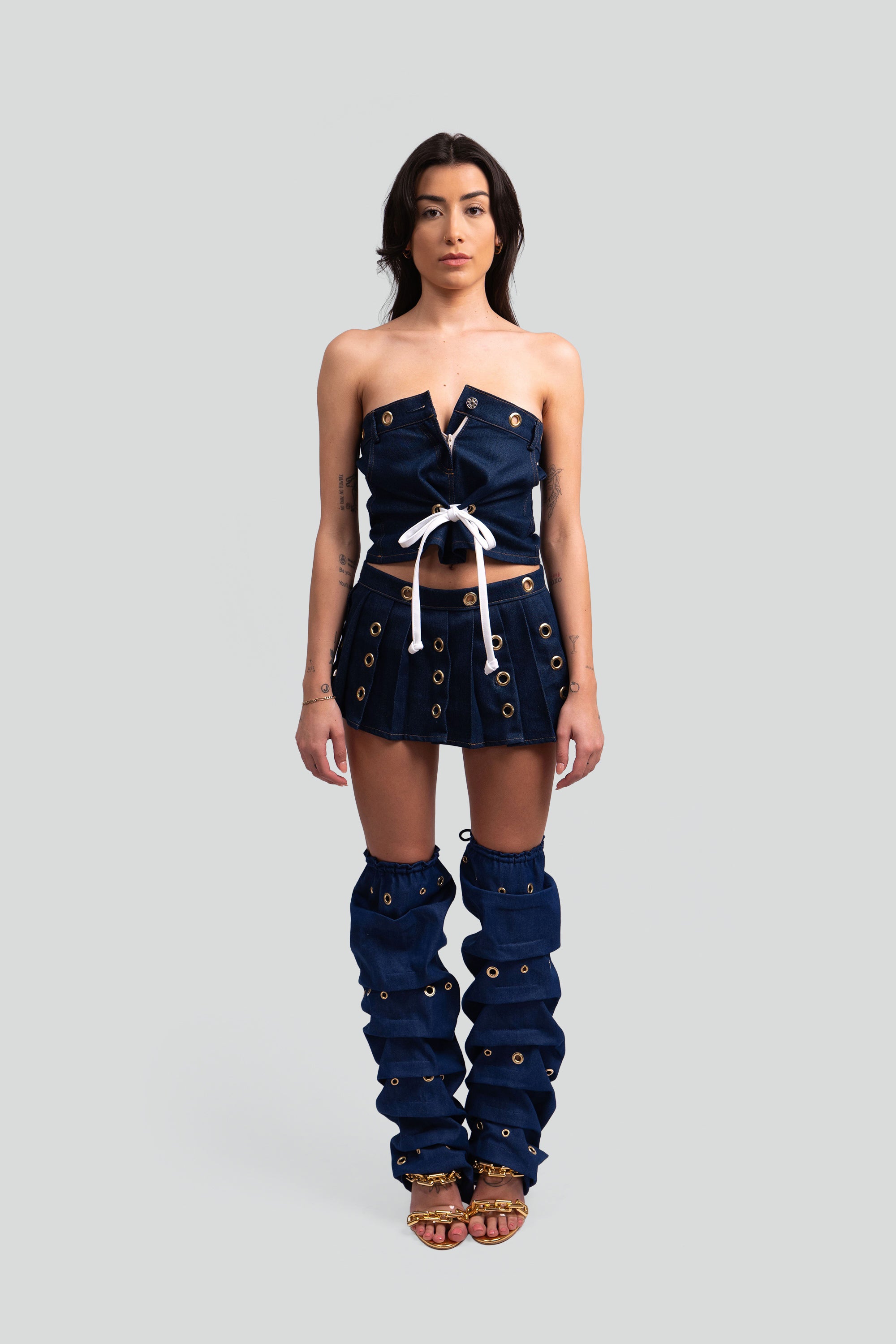 Woman wearing a denim outfit with a crop top, pleated skirt, and knee-high leg warmers with grommet details throughout on a white background