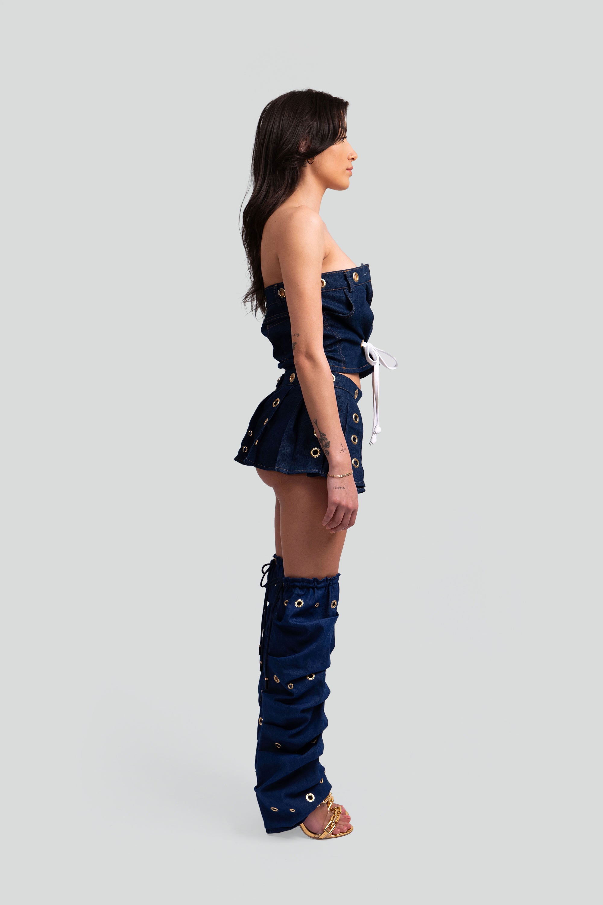 Side view of a woman wearing a denim outfit with a crop top, pleated skirt, and knee-high leg warmers with grommet details throughout on a white background