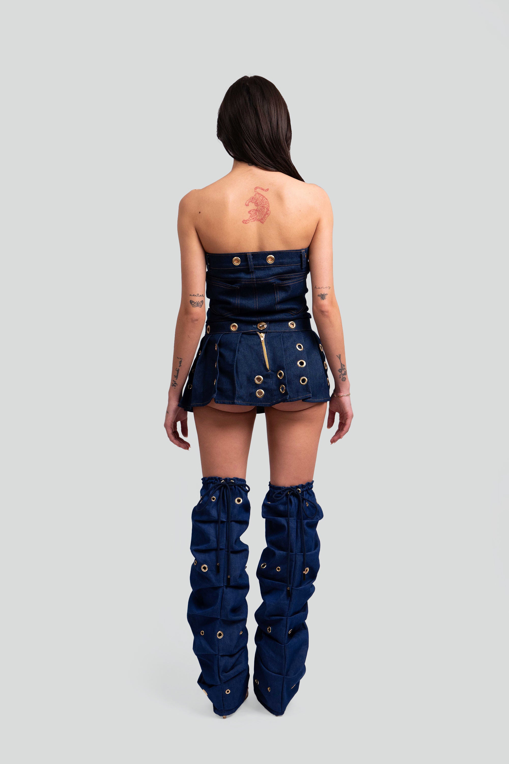 Back view of a woman wearing a denim crop top, pleated skirt, and knee-high leg warmers with grommet details throughout on a white background