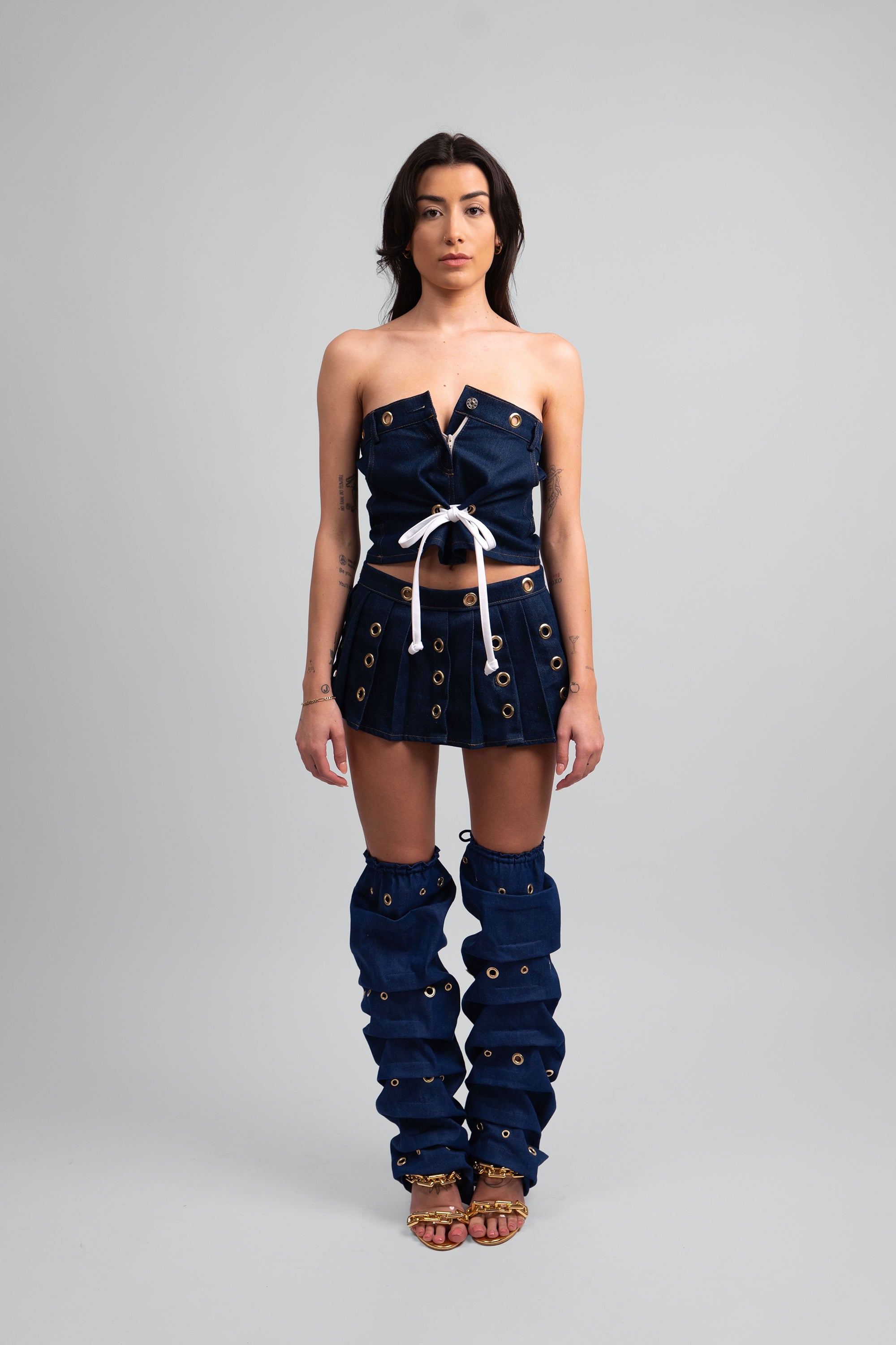 Woman wearing a denim outfit with a crop top, pleated skirt, and knee-high leg warmers with grommet details throughout on a white background