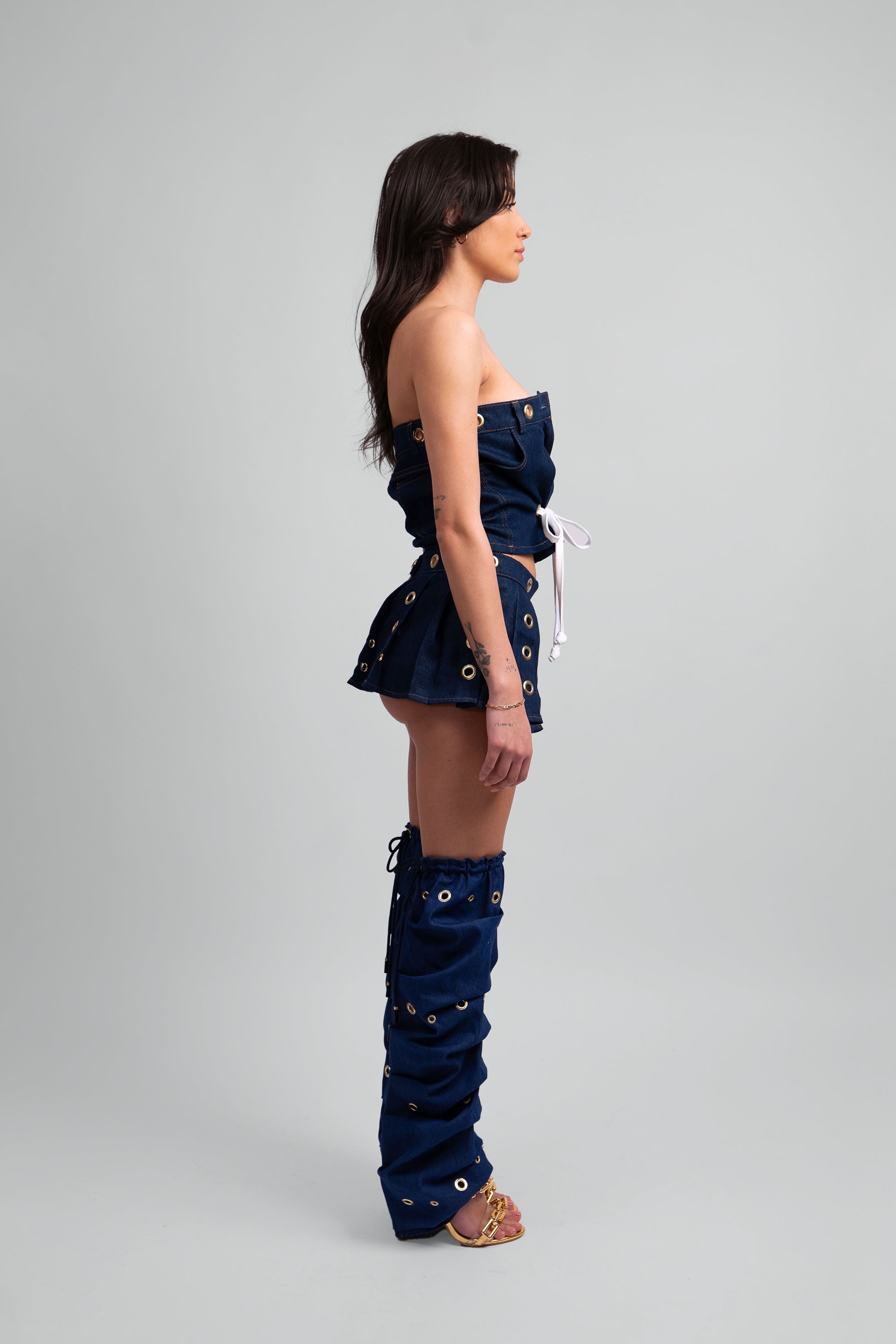 Side view of a woman wearing a denim outfit with a crop top, pleated skirt, and knee-high leg warmers with grommet details throughout on a white background