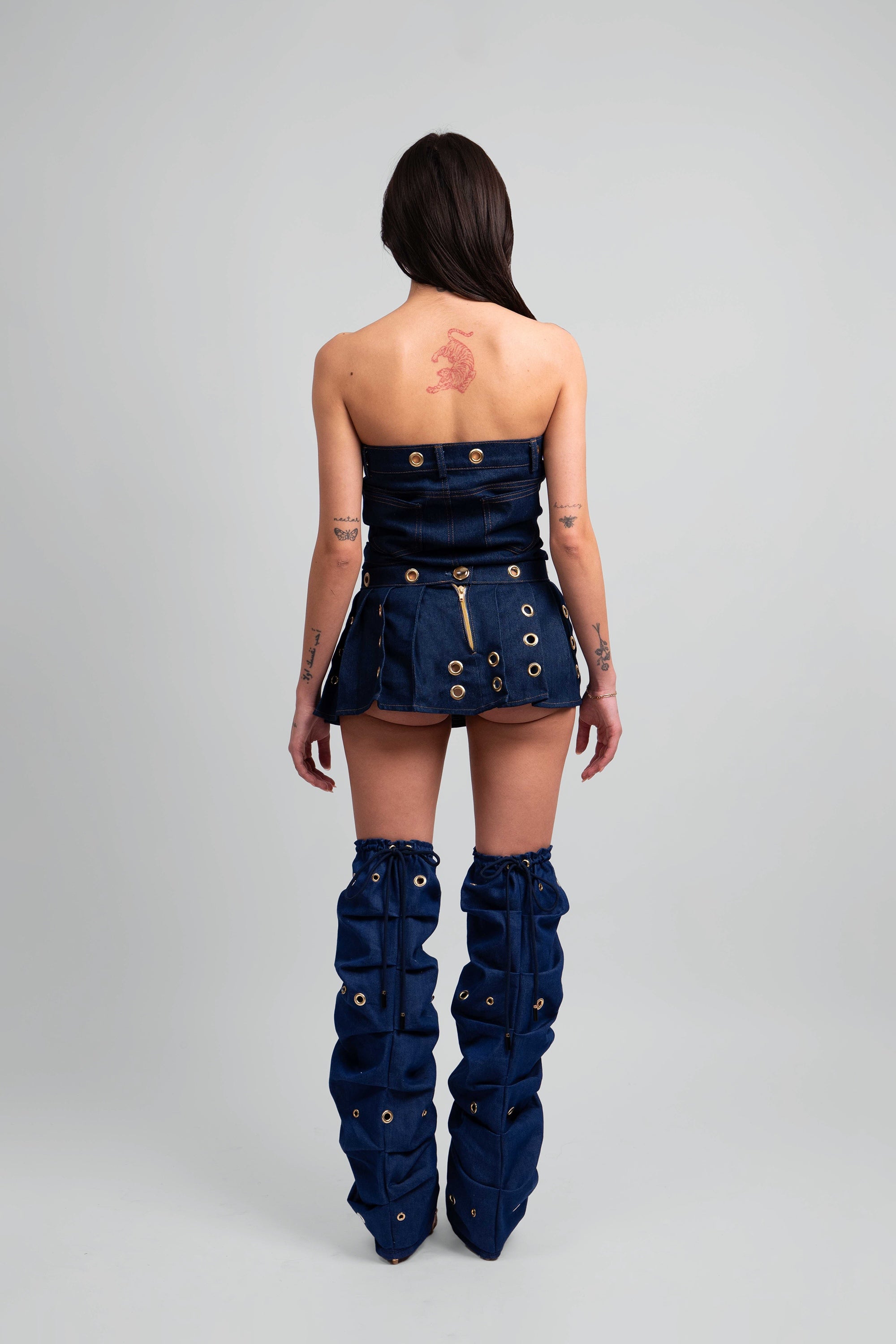 Back view of a woman wearing a denim crop top, pleated skirt, and knee-high leg warmers with grommet details throughout on a white background