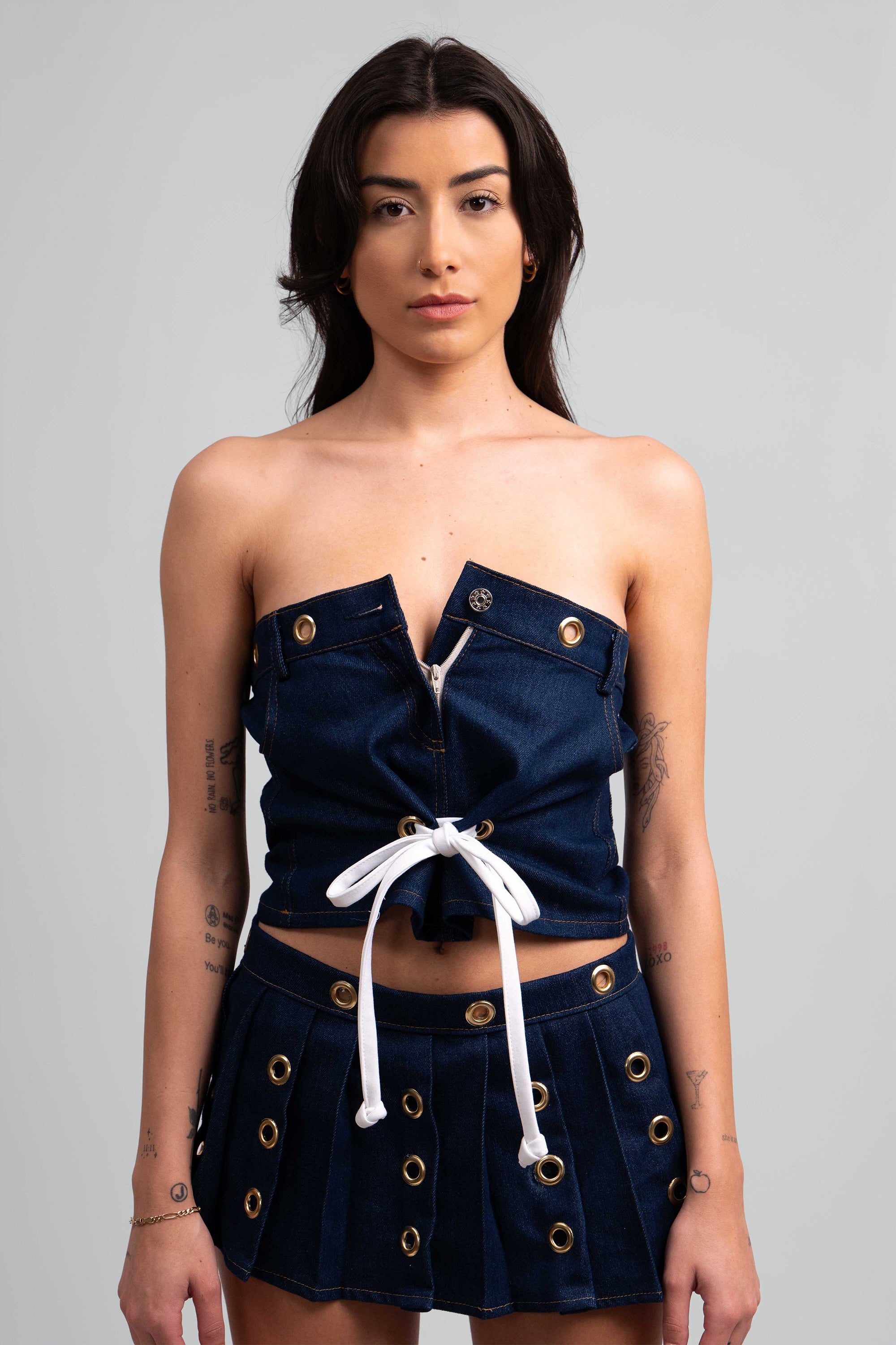 Close up of a woman wearing a denim outfit with a crop top and pleated skirt with grommet details throughout on a white background