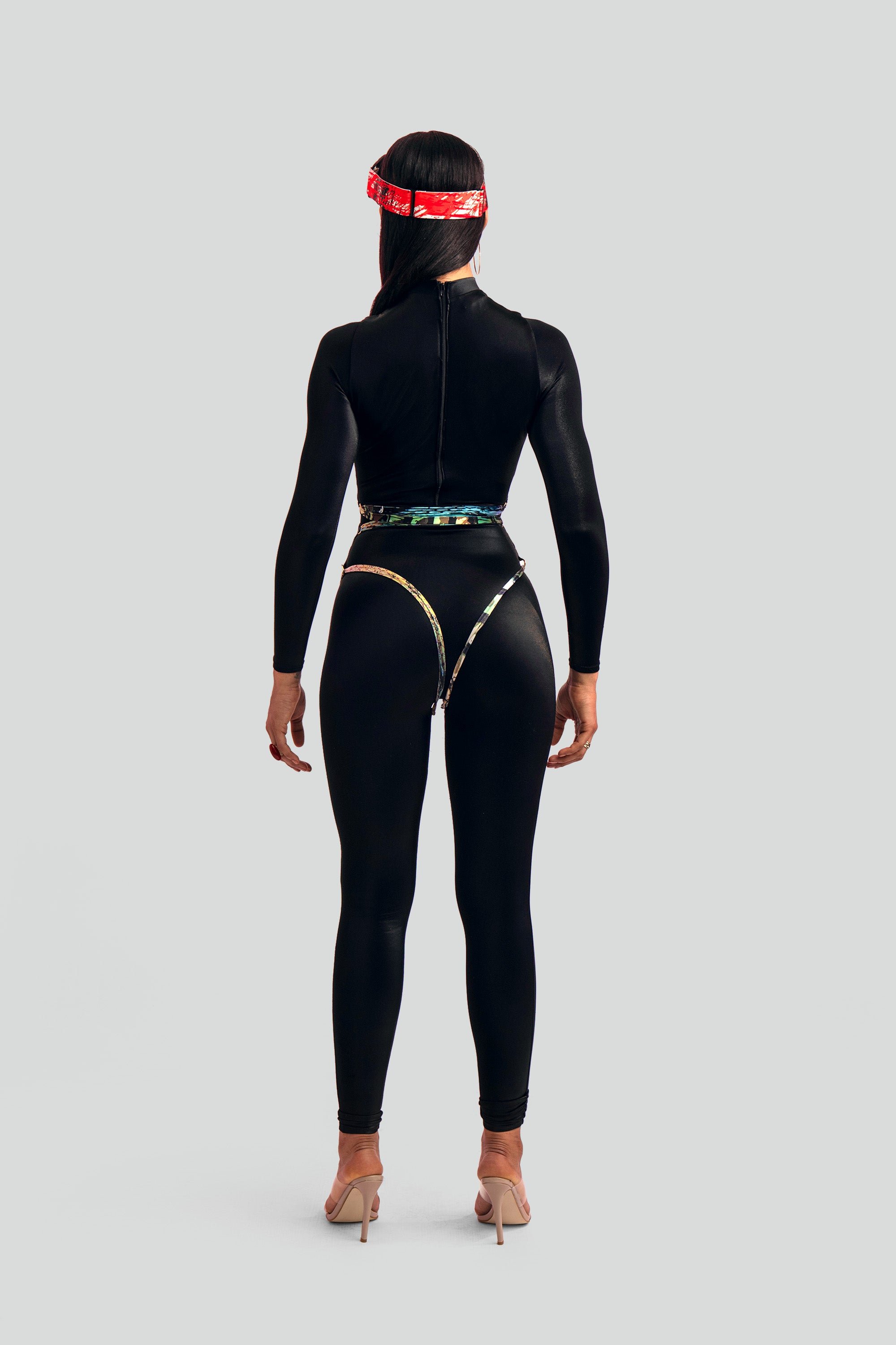 Back profile of a model wearing a sleek black catsuit highlighting gold rings along the sides and a multi-colored printed wrapping strap