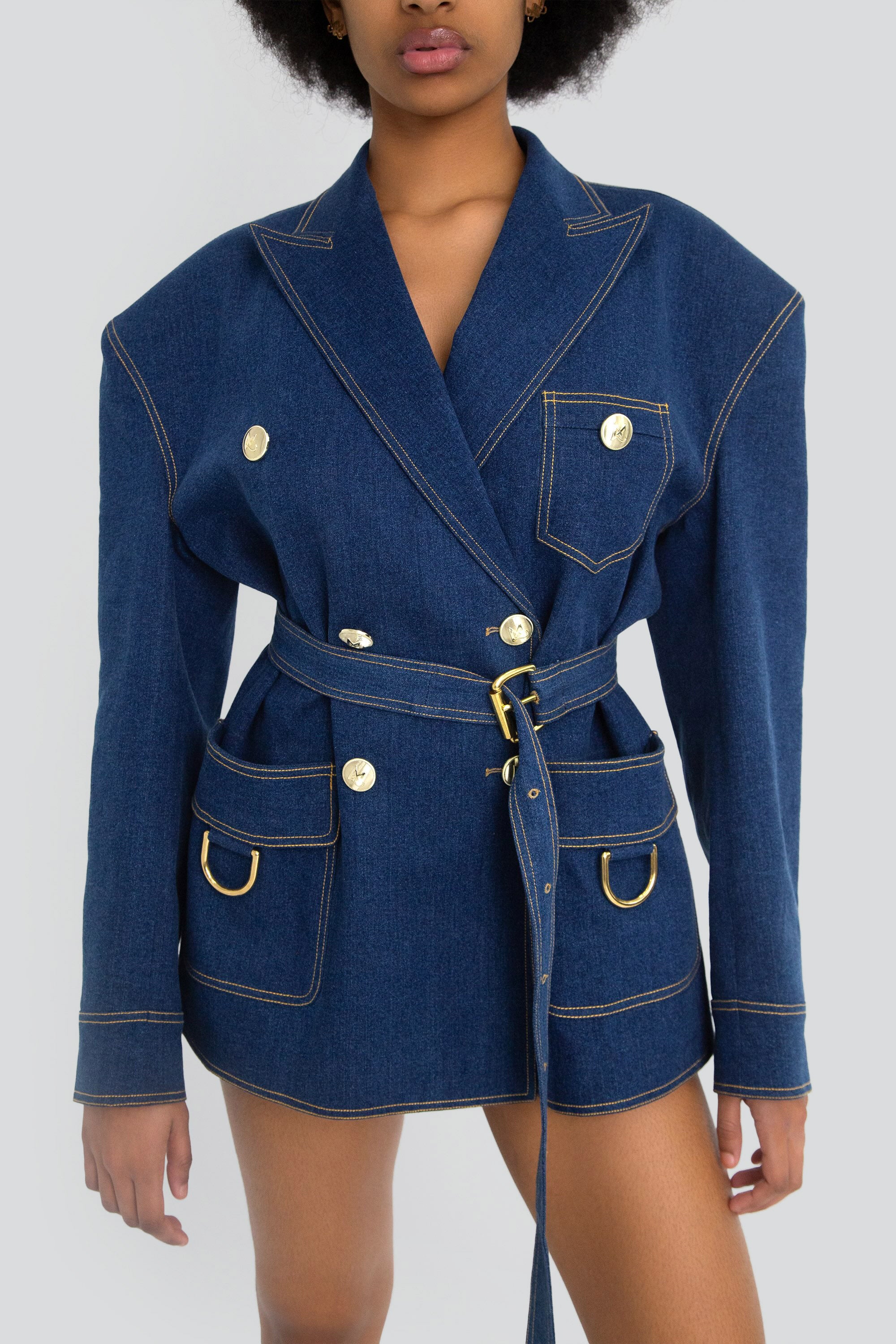 Woman wearing a blue denim blazer with gold hardware on a plain background