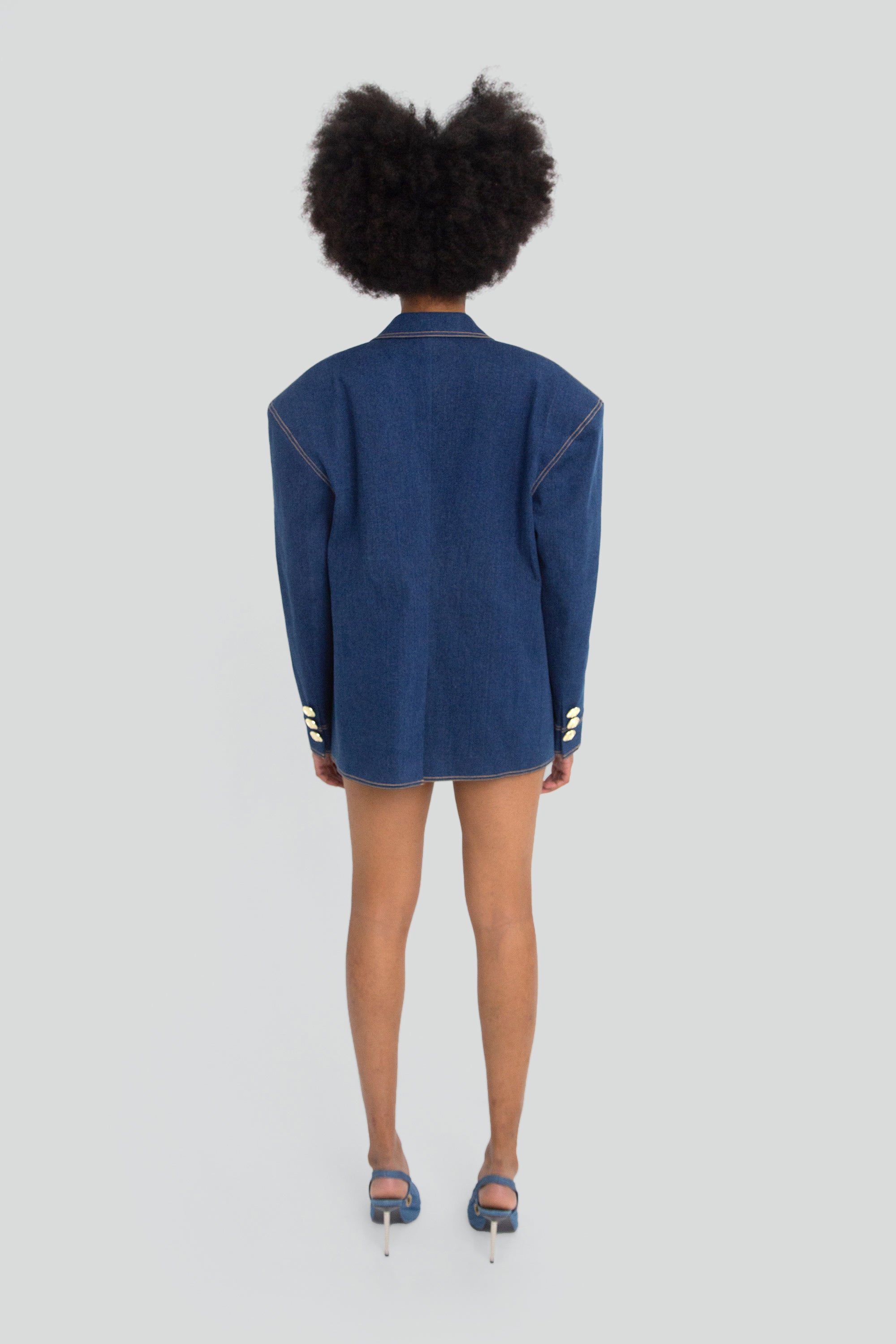 Woman wearing a blue denim blazer with gold hardware on a plain background