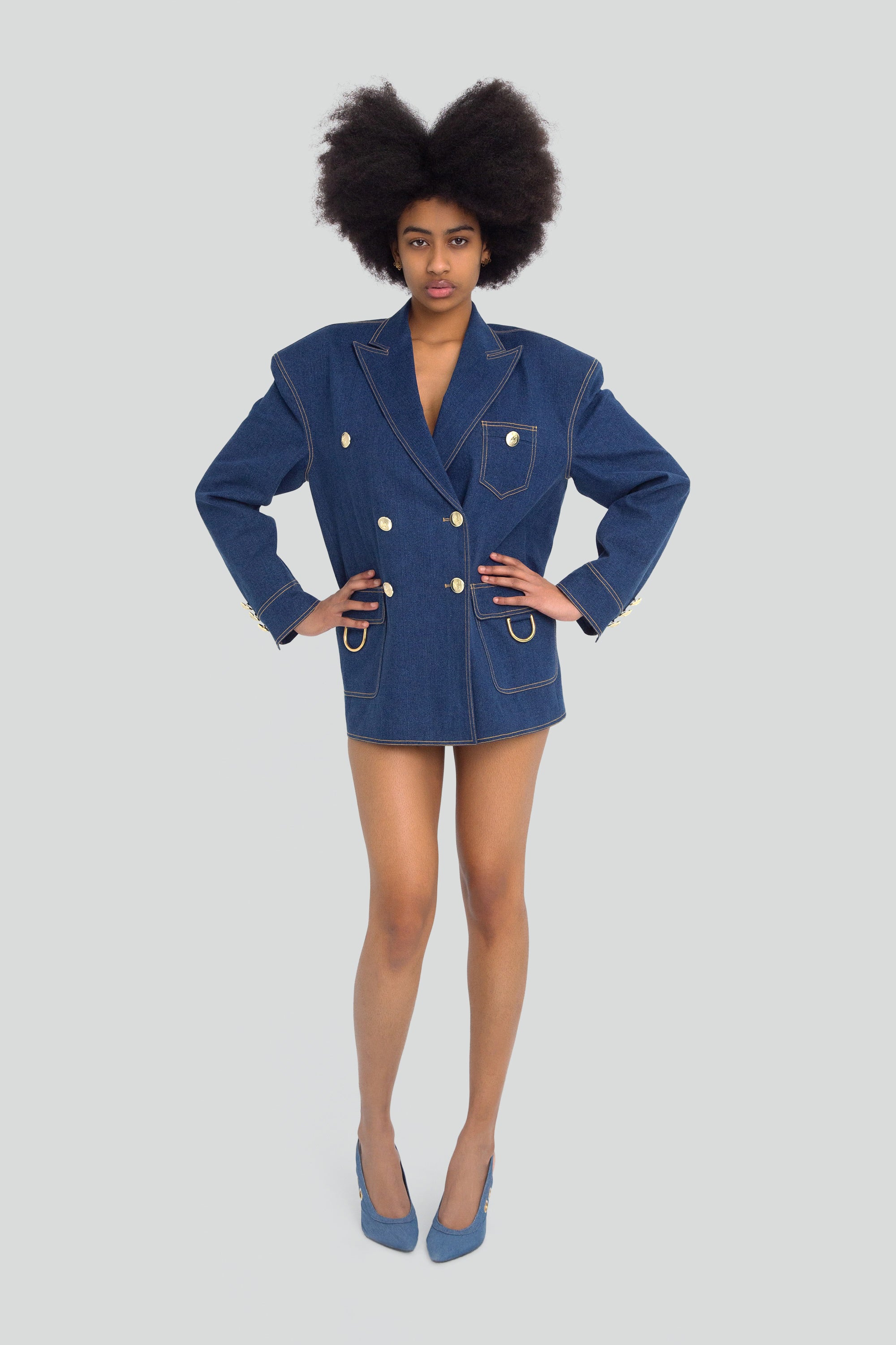 Woman wearing a blue denim blazer with gold hardware on a plain background