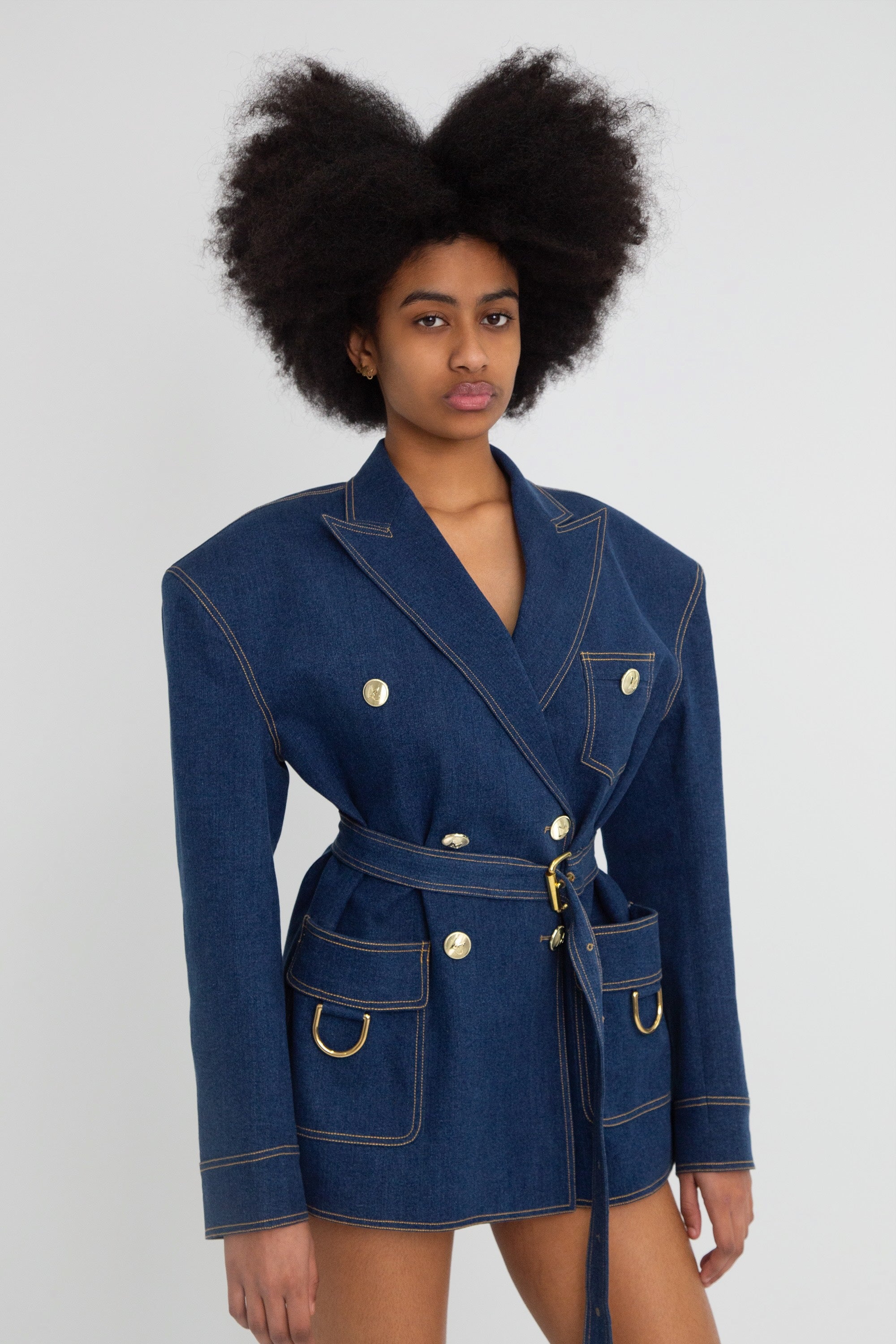 Woman wearing a blue denim blazer with gold hardware on a plain background