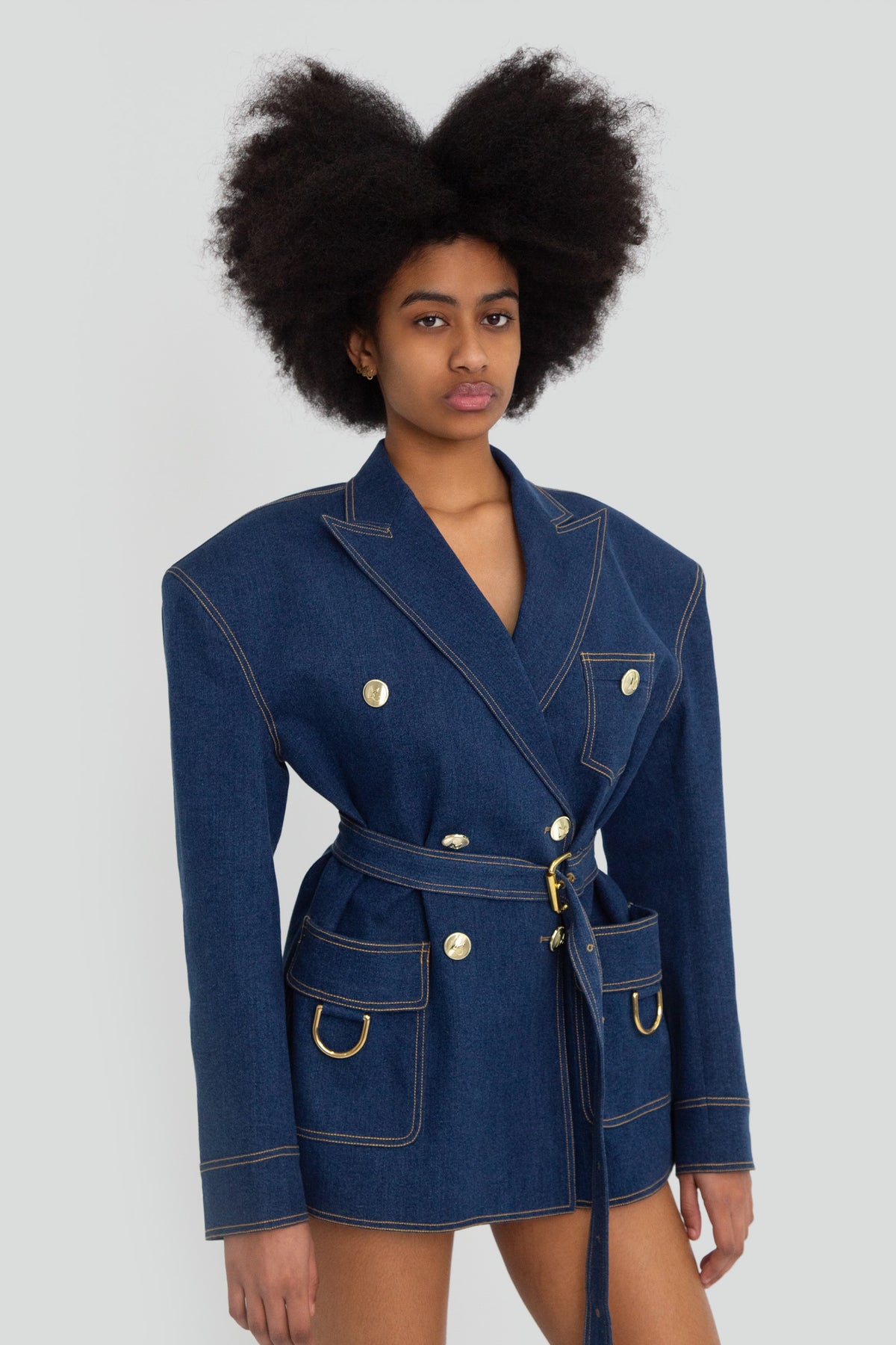 Woman wearing a medium blue denim box blazer jacket featuring structured padded shoulders, gold button details, and a removable waist belt