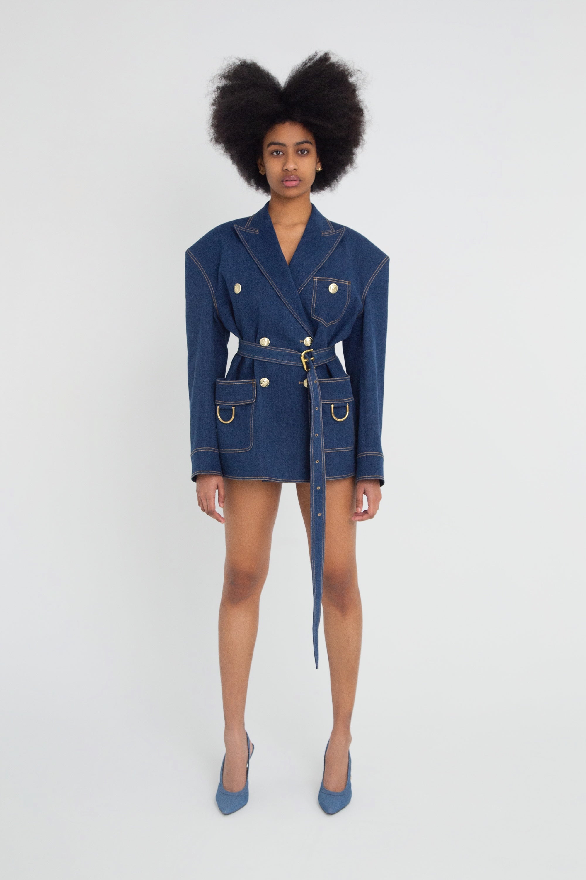 Woman wearing a blue denim blazer with gold hardware on a plain background