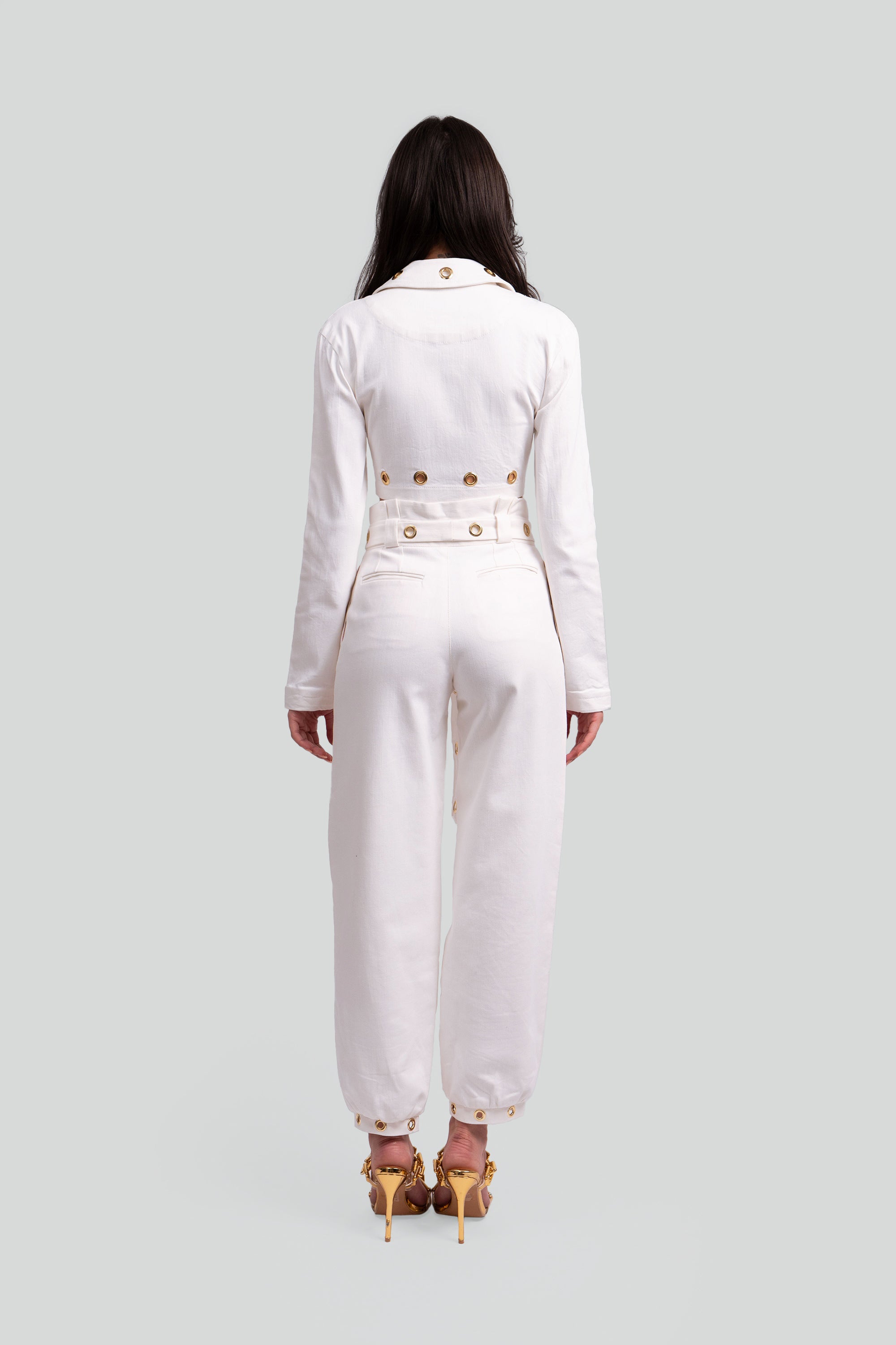 Back view of a woman wearing a white two piece cropped set with gold grommet details on a gray background